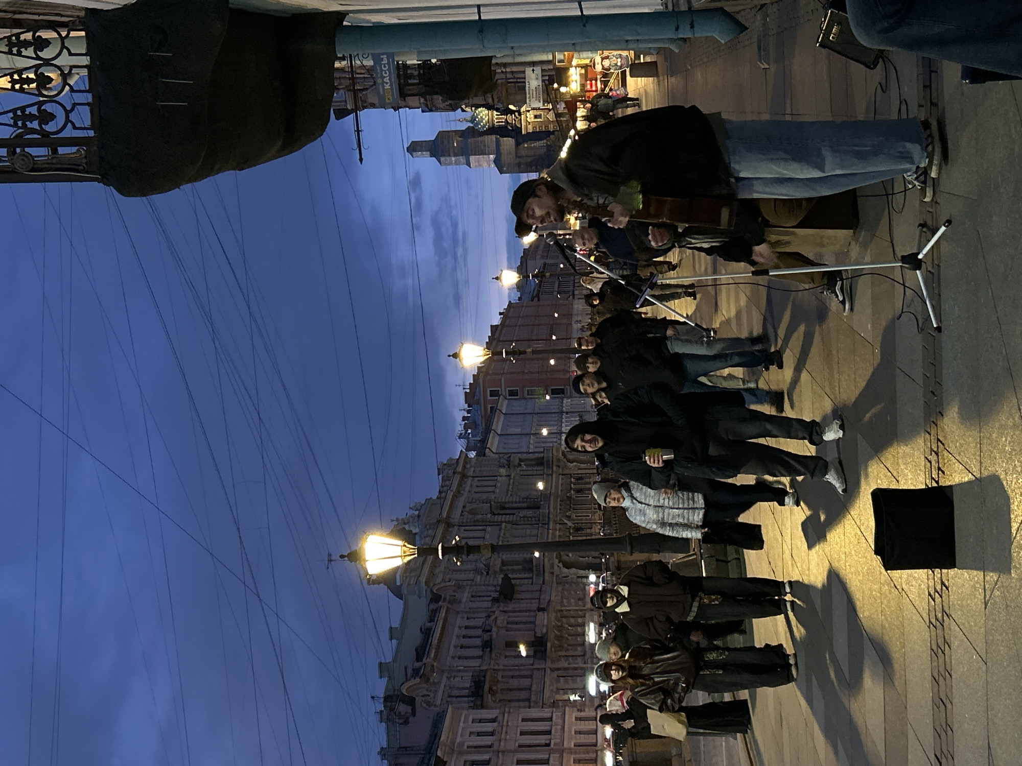 An evening street scene in Saint Petersburg, Russia, captures a street musician performing for a gathered audience. The sky is a deep twilight blue, illuminated by bright streetlights and the warm glow from buildings, signaling either dusk or early night.
The central figure is a male musician, wearing a dark cap and jacket, playing an acoustic guitar. He stands facing the audience, with a microphone stand and an amplifier positioned nearby, suggesting an amplified performance. A diverse group of people, casually dressed, are clustered around him, attentively watching and listening. Some audience members are holding cups, and their collective posture indicates engagement with the music.
In the background, ornate historic buildings with multiple stories line the street, their architectural details partially visible in the low light. Distant domed structures and other city architecture can be discerned against the horizon. Numerous thin power lines stretch across the dark sky. A prominent streetlight with a decorative fixture stands to the left, casting a strong, warm light onto the pavement and nearby audience members, creating long shadows.
A black case or bag rests on the ground in the foreground. On a sign visible on a building in the background, the Russian word "КАССЫ" (KASSY), meaning "Ticket Offices," is legible. The scene conveys a lively urban atmosphere, characteristic of a cultural street performance.