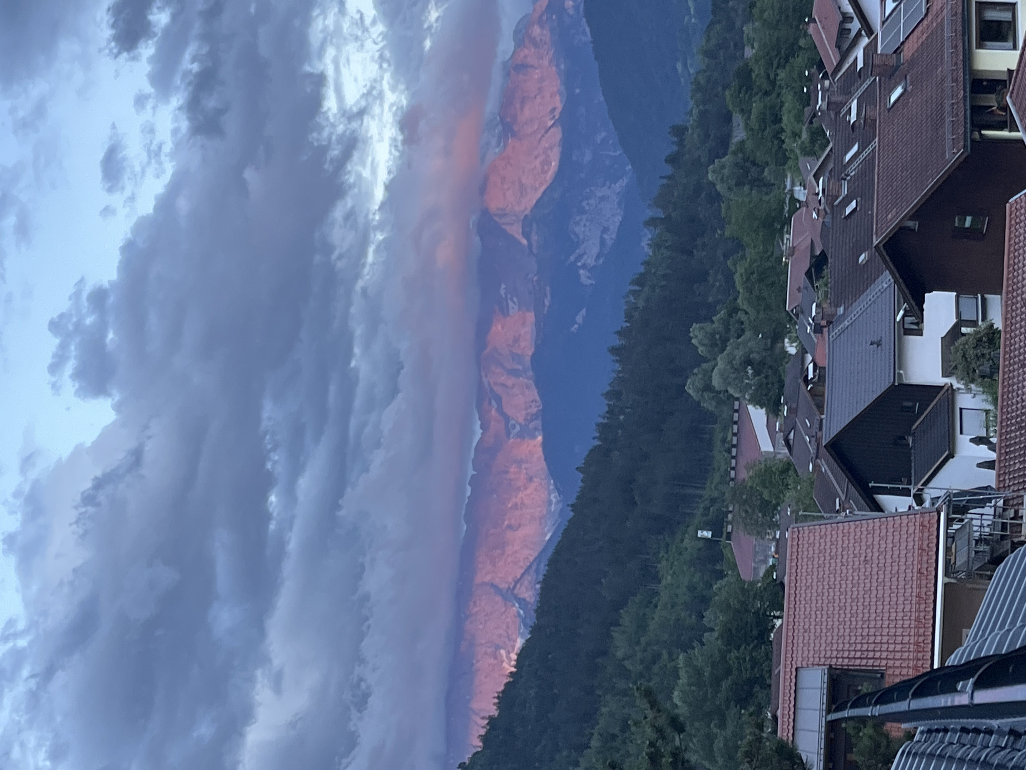The image captures a twilight view of the Zugspitze massif, Germany's highest peak, as seen from Garmisch-Partenkirchen. The rugged mountain range is illuminated by the setting sun, casting a warm pink and orange glow on its upper slopes. Dark, dramatic clouds gather in the sky, partially obscuring the higher peaks. In the foreground, the rooftops of Garmisch-Partenkirchen's houses are visible, interspersed with lush green trees. The scene conveys a sense of peaceful twilight in a mountainous Bavarian town.