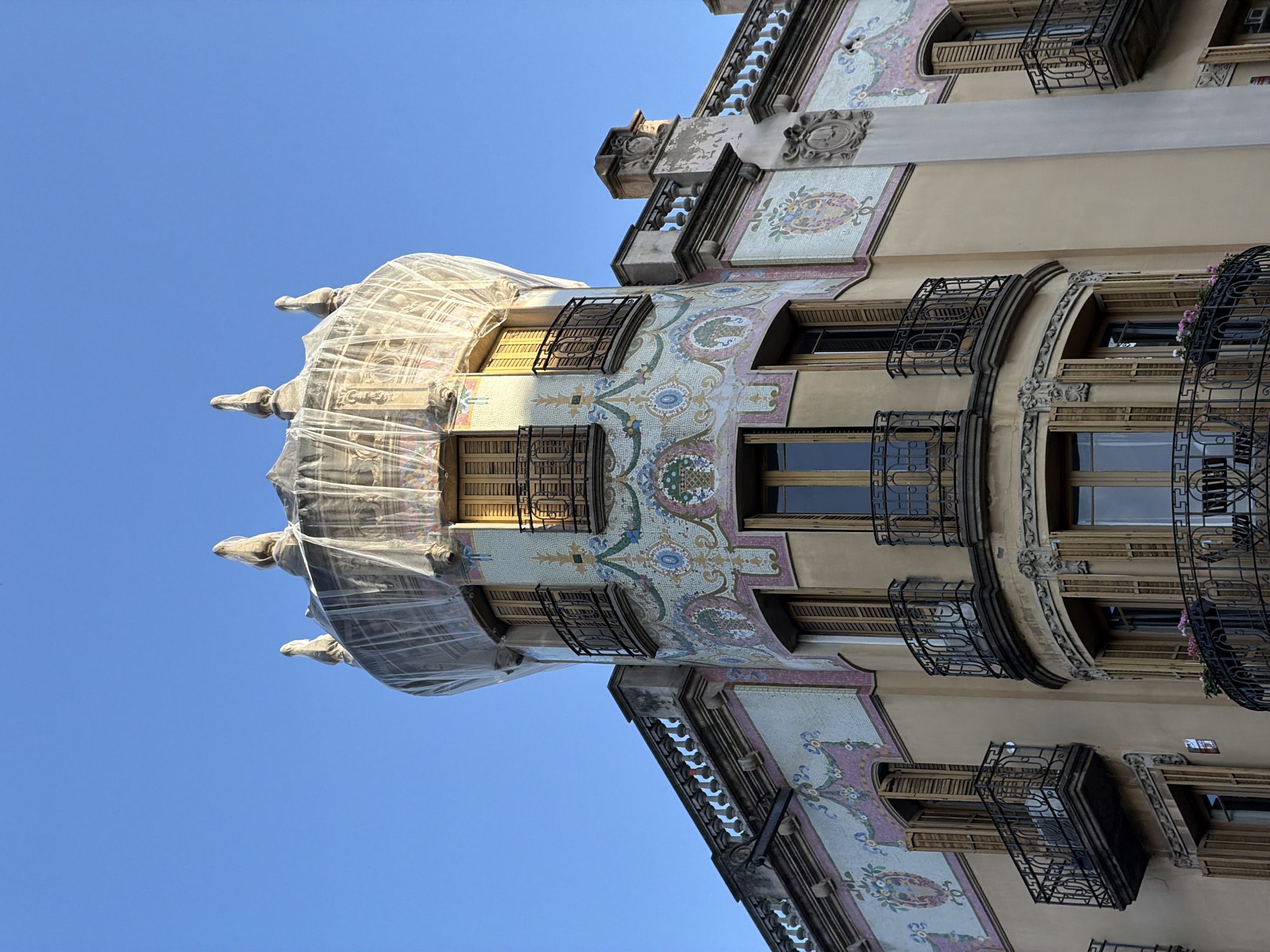 This upward-angled photograph captures a section of an ornate, multi-story building against a clear, vibrant blue sky. The architectural style, typical of Barcelona's Modernisme, is characterized by its curved facades and extensive use of colorful mosaic tiles, known as trencadís. These mosaics feature intricate floral and geometric patterns in shades of blue, green, pink, and cream.
The building is adorned with numerous windows fitted with wooden shutters and protected by decorative wrought iron grilles and balconies, many showcasing elaborate designs. A prominent turret-like section at the top of the building is partially shrouded in a translucent white mesh or netting, indicating ongoing maintenance, restoration, or protection of the structure. Beneath this netting, small, sculpted figures resembling gargoyles or decorative supports are visible. Further down, decorative balustrades and carved stone elements add to the building's intricate detailing.
The clear sky and bright light suggest it is daytime and sunny. No human activity is visible, focusing solely on the architectural grandeur. Faint, worn text can be partially discerned on the stone facade beneath the netting, appearing to read "GARAGE" followed by another word that is less clear, possibly "MOLINA" or "MORINA," suggesting a past business or historical detail.