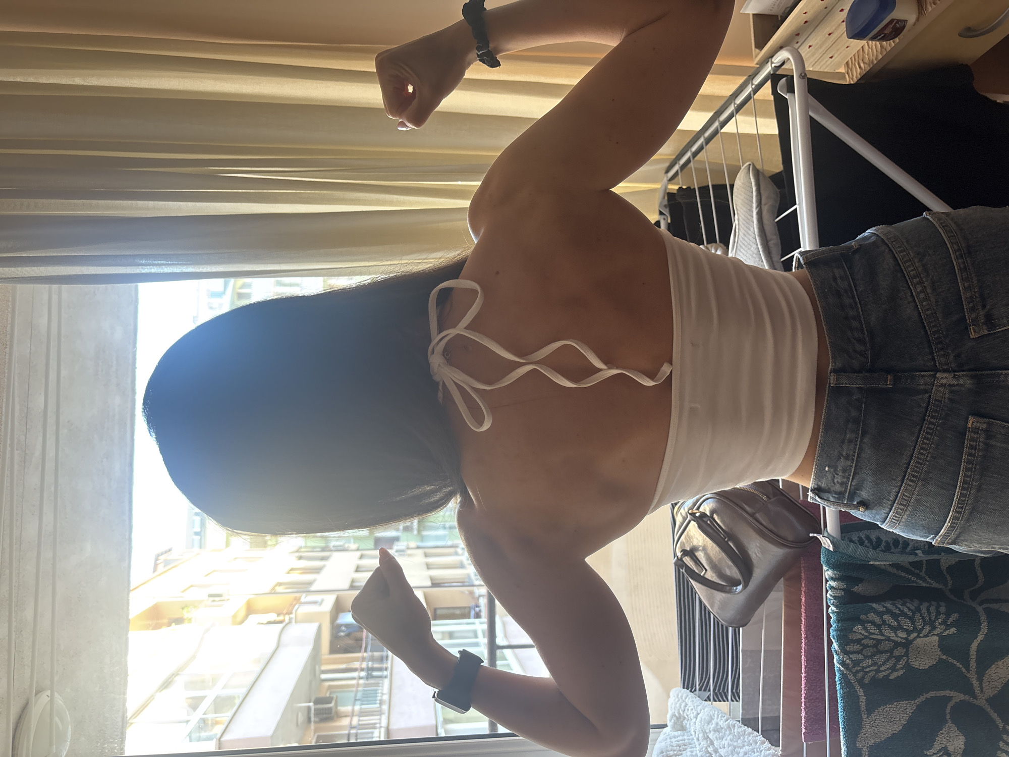 The image shows a woman from behind, wearing a white halter top and blue jeans, flexing her arm muscles. She is standing in front of a window with beige curtains, looking out at a city street in Varna, Bulgaria. The buildings across the street appear to be residential apartments, with balconies and air conditioning units visible. Sunlight streams through the window, illuminating the woman's back and creating a lens flare effect. In the foreground, there is a clothes drying rack with various items of laundry, a handbag, and a small jar of Vaseline. The scene suggests a domestic setting within a city environment.