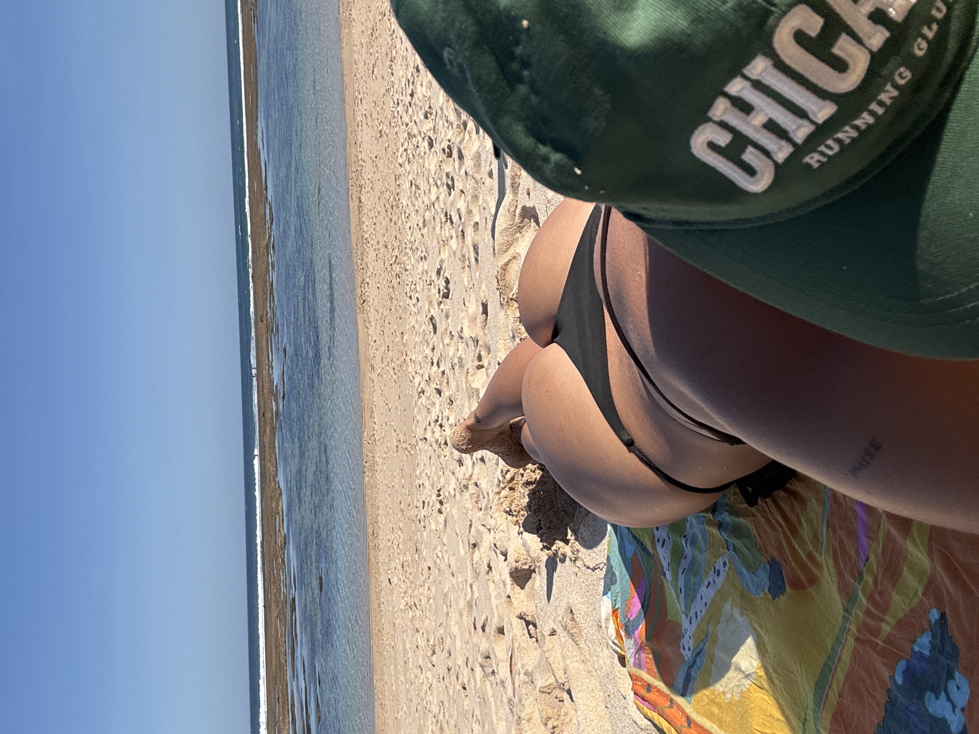 A person wearing a black bikini is lying on a beach on a colorful patterned blanket. The person is wearing a green baseball cap that reads "CHICAGO RUNNING CLUB". The scene is an outdoor beach with sand and a view of the ocean and blue sky. The location is Kuta Selatan, Indonesia.
