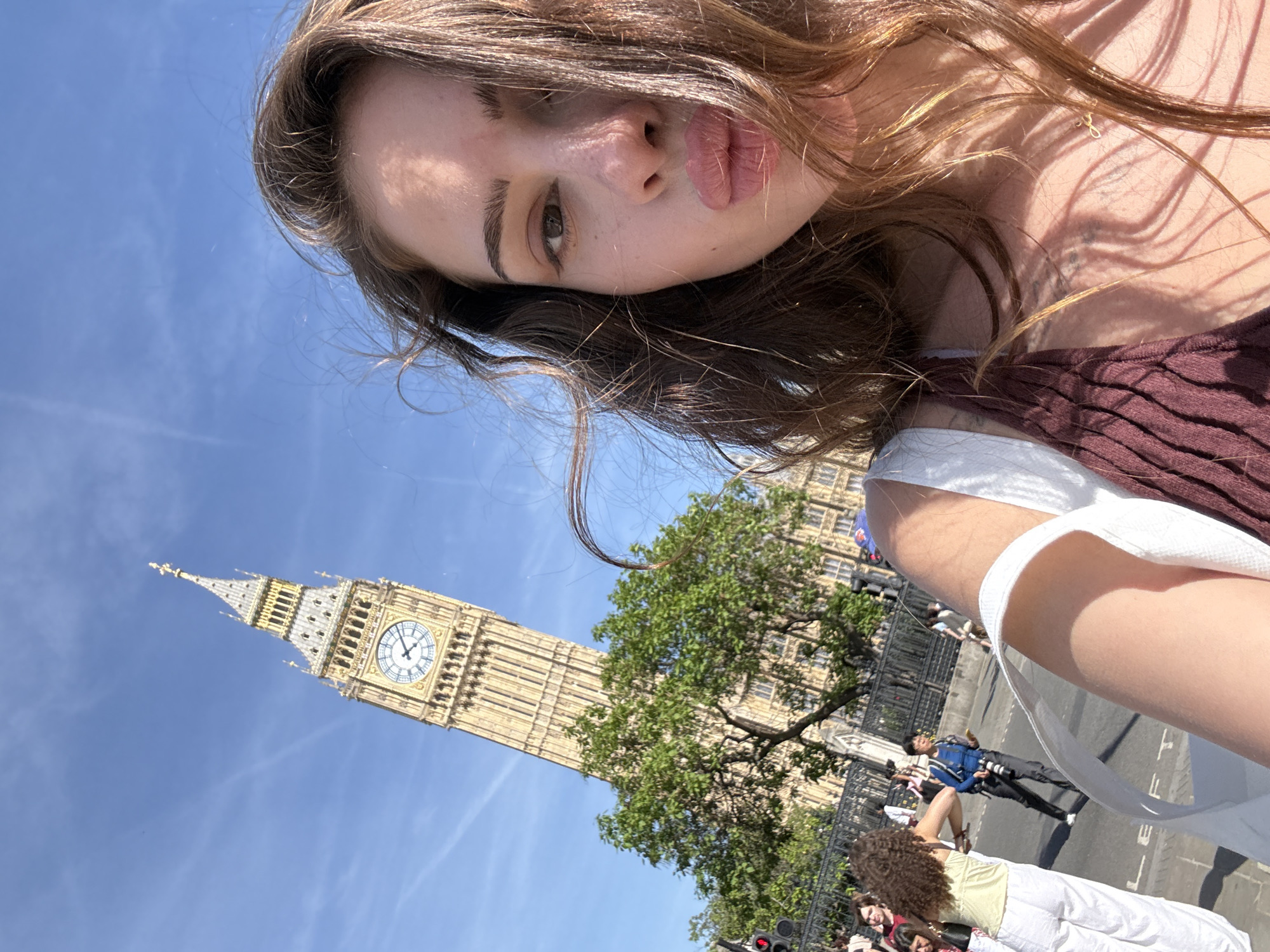 A selfie of a woman with long brown hair in front of the Elizabeth Tower, also known as Big Ben, in London, United Kingdom. She is pouting her lips at the camera. The tower is visible to the left of her, with its clock face and architectural details. A tree partially obscures the tower's base. The background shows a street with pedestrians and a red traffic light. The sky is blue with light clouds. The woman is wearing a burgundy top and a white strap.
