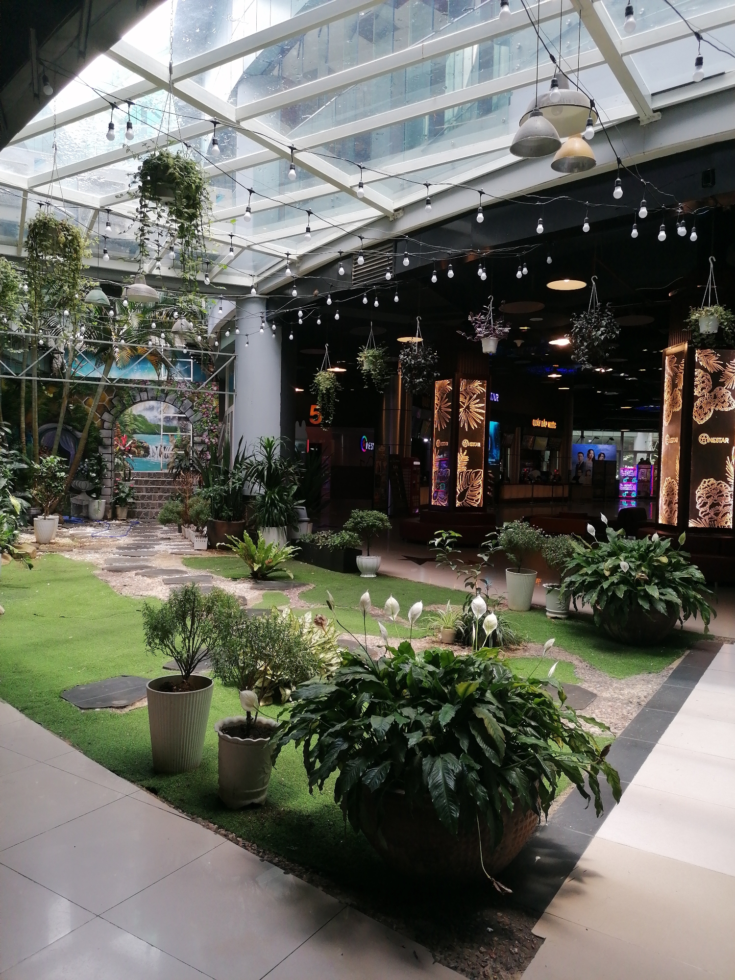 The image depicts an indoor garden within a shopping center in Da Lat, Vietnam. The garden features artificial grass, stepping stones, and various potted plants, including peace lilies and ferns. The backdrop of the garden is a mural depicting a tropical landscape with a waterfall. The garden is illuminated by natural light from a glass ceiling, as well as decorative string lights and pendant lamps.
Beyond the garden is the interior of the shopping center, with storefronts, illuminated display panels, and a glimpse of people inside. The flooring in the shopping center transitions from artificial grass and gravel in the garden to tiled flooring. The building appears modern, with glass and steel construction.
