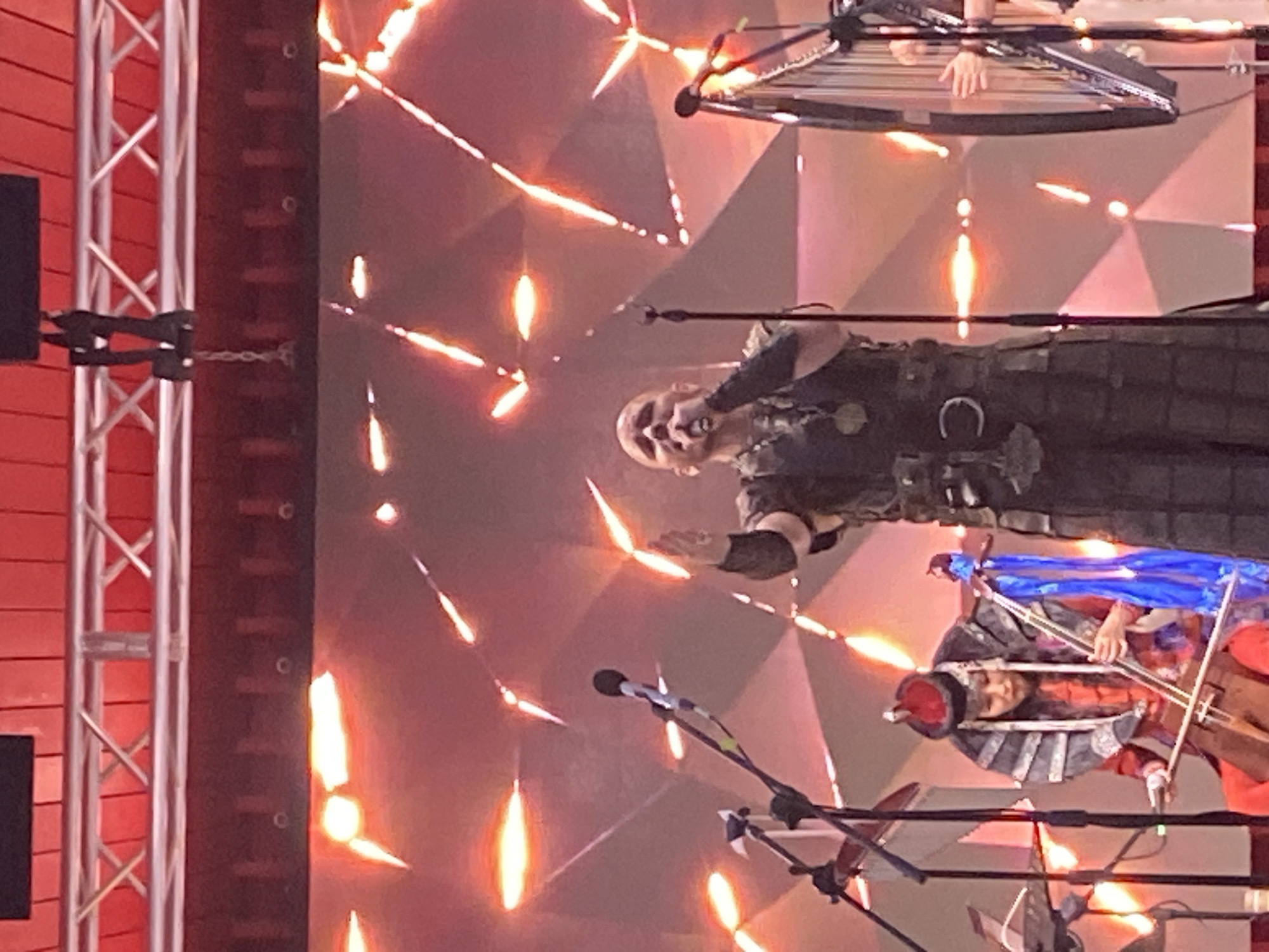 In Moscow, Russia, a band performs on a stage bathed in red and orange light. A male singer, bald and wearing a dark, ornate vest and pants, stands center stage, singing into a microphone, his right hand raised. To the left, a woman in traditional Mongolian attire, including a red hat and ornate collar, plays a two-stringed fiddle-like instrument. To the right, a harp stands partially visible, with a person's hands on its strings. Microphones and lighting rigs are visible throughout the scene. The backdrop is a large screen displaying a geometric, fiery pattern. The overall impression is of a live musical performance.