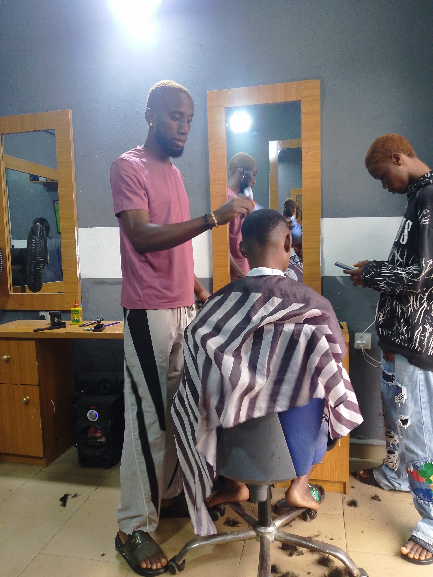 In a barber shop located in Agbani, Nigeria, three young men are present. One man, with blonde hair and wearing a pink t-shirt and grey pants, is standing and appears to be a barber. He is holding a tool, possibly scissors or a comb, and is in the process of cutting the hair of another young man. This individual is seated in a barber chair, covered with a striped cape, and is facing away from the camera. His hair is short. A third young man, also with blonde hair, is standing to the right of the seated customer, holding a smartphone and observing the haircut. He is wearing a black hoodie with white designs and distressed blue jeans. The barber shop environment features mirrors on the walls, a wooden counter with barber tools and a bottle of product, and a speaker system. The floor is tiled and has scattered hair clippings, indicating a recent haircut. The overall lighting suggests an indoor setting, possibly with artificial light sources.