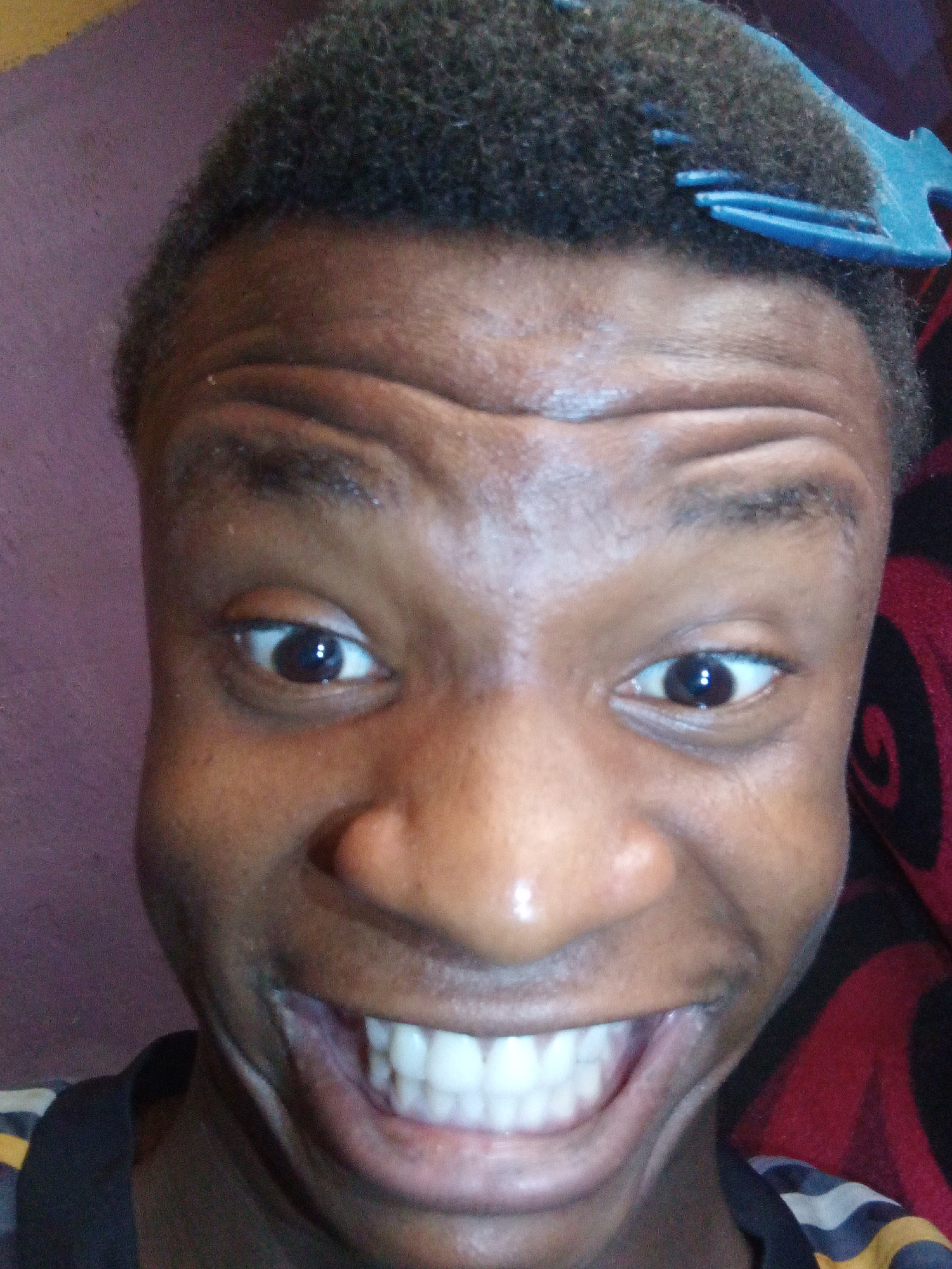 A close-up, distorted selfie of a Black male. He has short, dark, curly hair and a blue comb inserted near the front of his hairline. His forehead is heavily wrinkled. His eyes are open and looking directly at the camera. His mouth is wide open, displaying a full set of teeth. The background is a combination of a purple wall and a red and black patterned surface, possibly a blanket or cushion. The image was taken in Narayi, Nigeria.