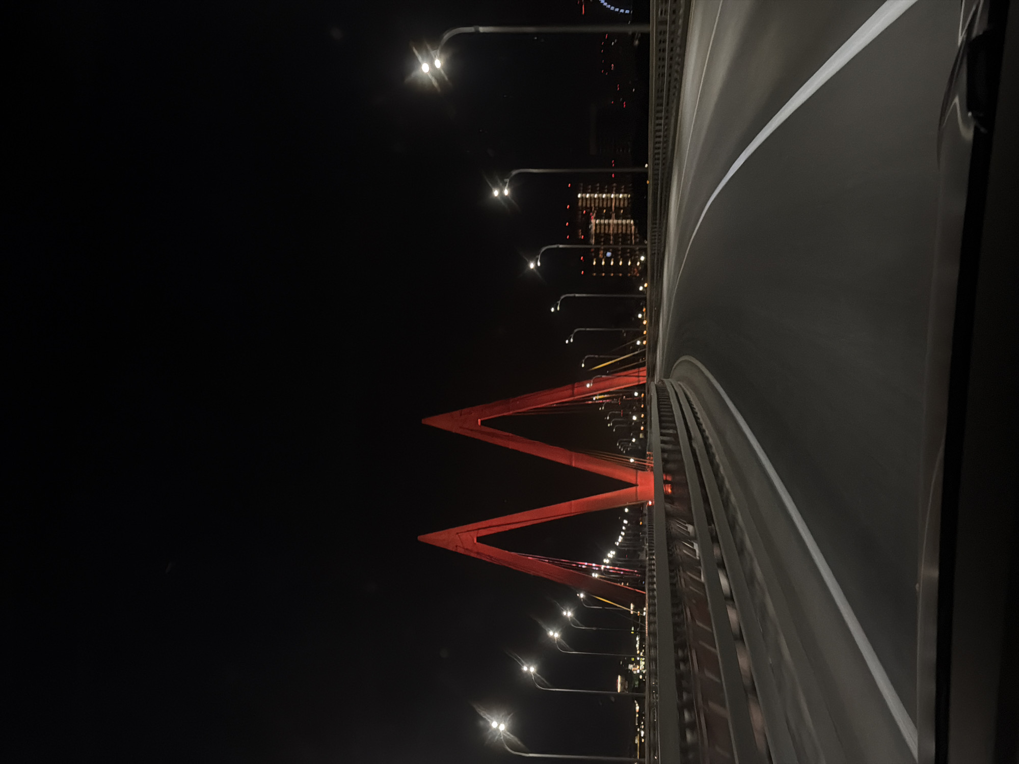 The image captures the Millennium Bridge in Kazan, Russia, at night. An empty road curves across the bridge, marked with white lines. The bridge is illuminated by streetlights lining its side. The red "M" shaped pylon of the bridge is prominently featured. In the background, city lights are visible. The environment is dark, with a clear night sky. There are no people visible in the frame. The camera angle is from a low perspective, emphasizing the bridge's structure.