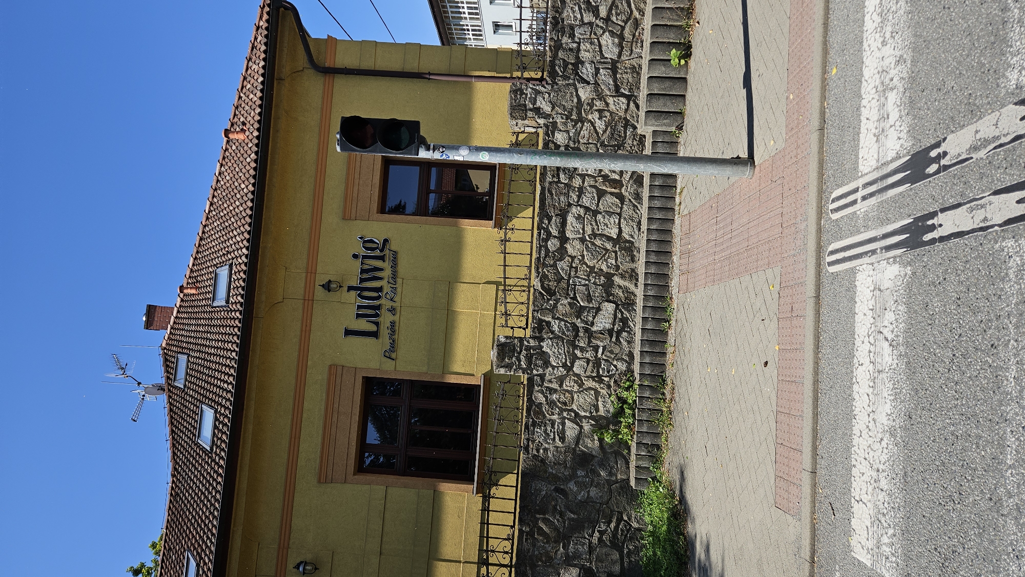 The image shows the building of a "Ludwig Penzión & Restaurant" located in Prešov, Slovakia. The building is yellow with a red tiled roof and several skylights. A wrought-iron fence separates the building from a sidewalk made of grey and red bricks. A stone wall is visible behind the fence. A traffic light stands on the sidewalk in front of the building. The foreground shows a crosswalk on a paved street. The sky is clear and blue, and the sun is shining, casting shadows. There are no people visible in the image.