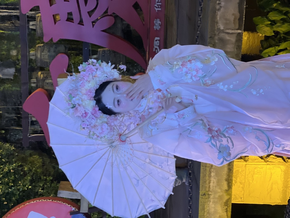 A female individual is centrally depicted in a night-time photograph taken in Yuzhong District, China. She wears a light pink traditional Chinese-style robe, embellished with intricate floral embroidery in pink, white, and teal on the sleeves and front. Her head is adorned with a large, elaborate floral headpiece composed primarily of pink and white blossoms. Her face features cosmetic white cross-shaped markings above her eyebrows and glitter on her cheekbones. She holds a white paper parasol with a wooden handle in her left hand, angled upwards and to her left. Her right hand is raised, partially covering her mouth. The background features large, red illuminated Chinese characters, prominently displaying "唐" (Tang) and smaller characters including "洞 梦 你" below it, indicating signage or decorative elements. Behind these, a series of dark, bell-like structures hang. The scene is illuminated by warm yellow-orange light originating from the lower left, highlighting a stone or concrete structure. Dark foliage and a section of water are visible towards the bottom right of the frame.