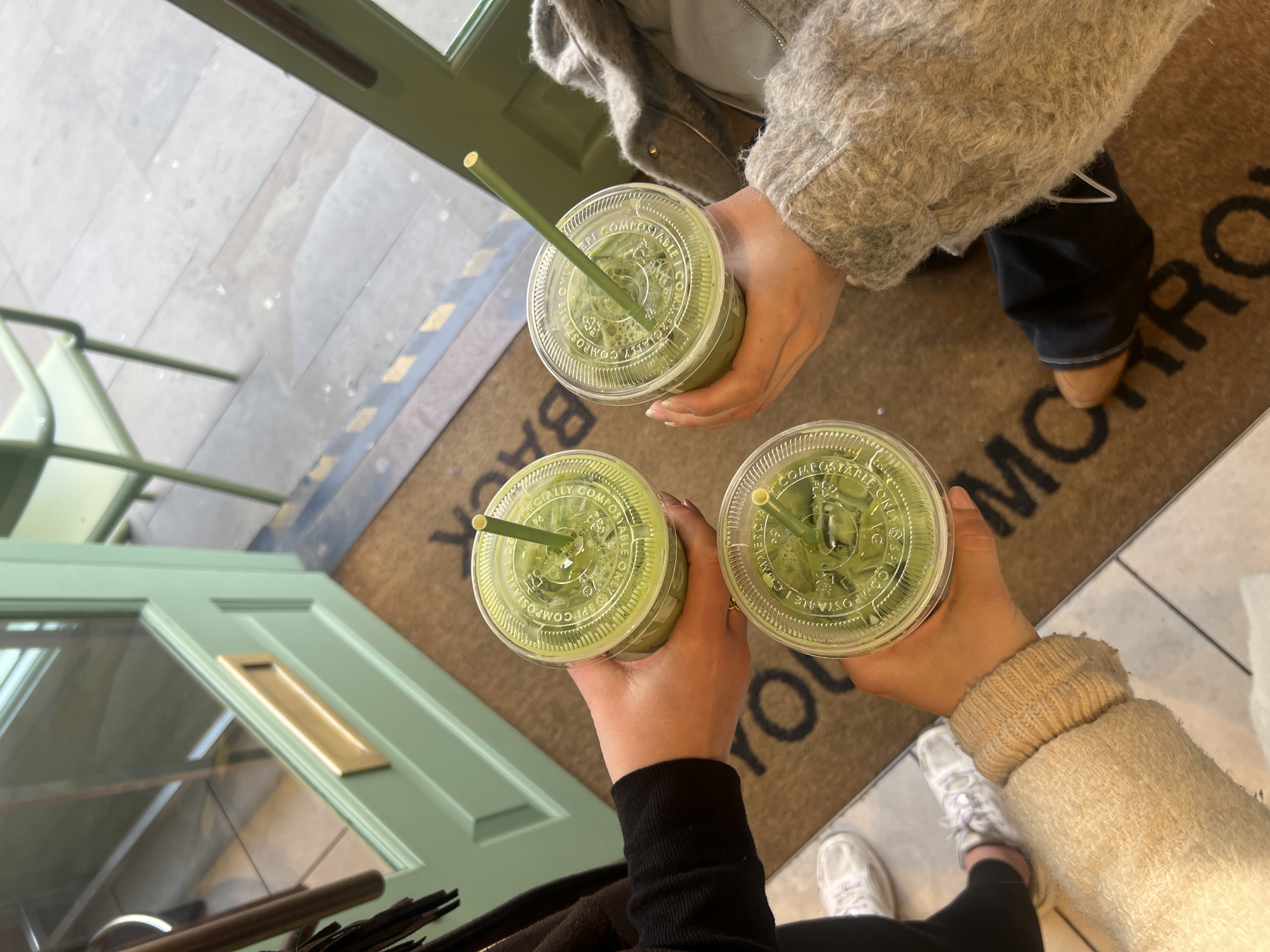 An overhead photograph captures three individuals holding clear plastic cups of a light green, frothy beverage, each with a green straw, converged in a central gesture. The plastic lids of the cups are embossed with the word "COMPOSTABLE." One person, positioned on the left, wears a black long-sleeved top and white athletic shoes. Another, on the right, wears a grey textured, possibly wool or fleece, long-sleeved jacket. The third, at the bottom of the frame, wears a light beige ribbed long-sleeved garment and partially visible brown shoes. The scene is located at the entrance of a business in the city of London, United Kingdom, specifically identified by a brown doormat with the word "TOMORROW" printed in black capital letters. The entrance features a partially open, light green wooden door with a gold-colored letterbox. Beyond the door, an outdoor area is visible with a light green metal chair and grey paved flooring. A yellow and black striped safety marker is present on the floor at the threshold.