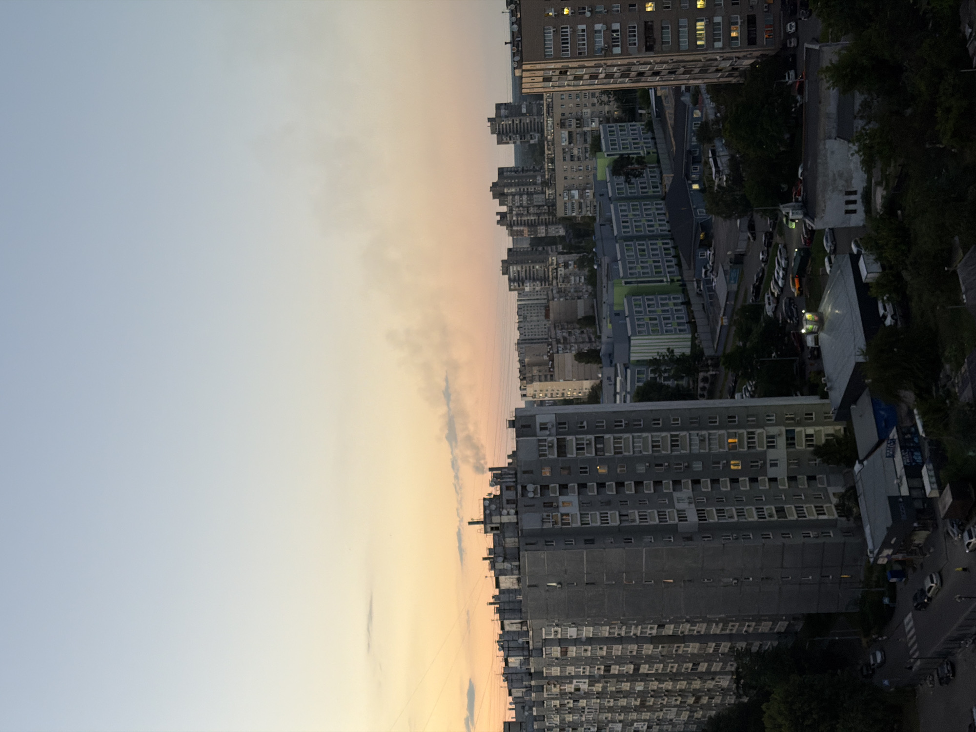 The image depicts an urban landscape at dusk, likely in Kyiv, Ukraine, given the architectural style of the buildings. The sky transitions from a soft orange hue near the horizon to a pale blue at the top. Wisps of smoke are visible rising from one of the taller apartment buildings on the right side of the frame.
In the foreground, a series of multi-story apartment buildings dominate the scene. The building on the left is a tall, grey concrete structure with numerous balconies and visible windows, some of which are illuminated. Adjacent to it, to the right, is another similar building. Further into the background, more apartment blocks of varying heights and architectural designs are visible, some with green facades.
A street level view shows cars parked along the side of the road and in parking lots. Pedestrians are present, including a person in yellow clothing near the street. Several smaller commercial or industrial-looking buildings are situated at the base of the apartment blocks. The overall impression is of a densely populated residential area during the evening hours, with hints of industrial or domestic activity suggested by the smoke.