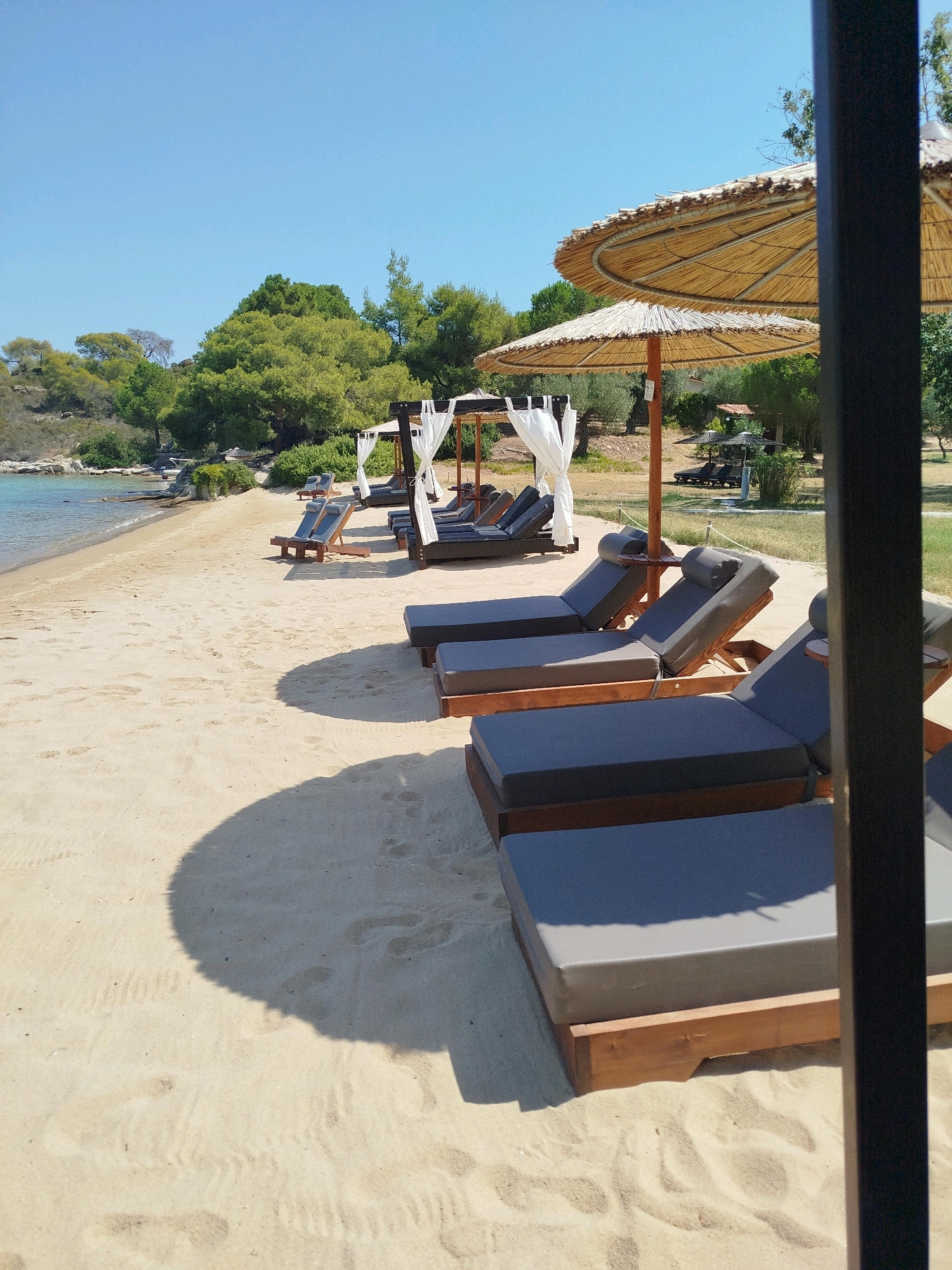 This image captures a serene beach scene in Sithonia Municipal Unit, Greece, under a clear blue sky. In the foreground, several dark grey cushioned lounge chairs are arranged on a sandy beach. Some of these chairs are under thatched umbrellas, providing shade. The chairs are constructed with wooden frames, and some have small wooden tables attached. In the background, a calm body of turquoise water meets the shore. Beyond the beach, a lush green hillside is covered with trees, suggesting a natural, unspoiled environment. Further back, there are a few more lounge chairs and umbrellas, indicating a beach resort or a well-maintained public beach. The overall impression is one of a peaceful and inviting vacation destination.