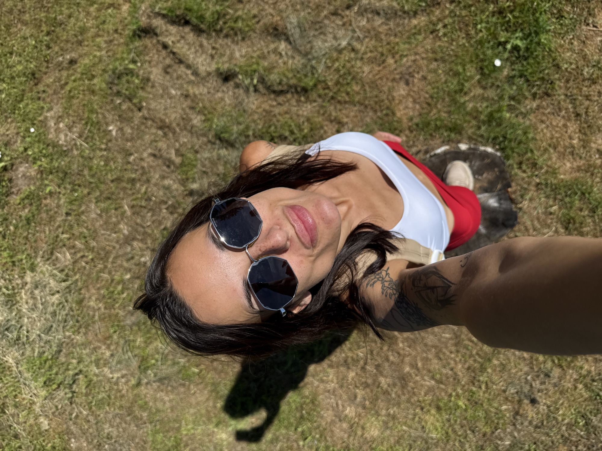 A woman is standing outdoors, seen from a high angle. She is wearing sunglasses, a white top, and red pants. She has tattoos on her arm. The ground is covered in grass and dry vegetation. The sun is casting a shadow on the ground.