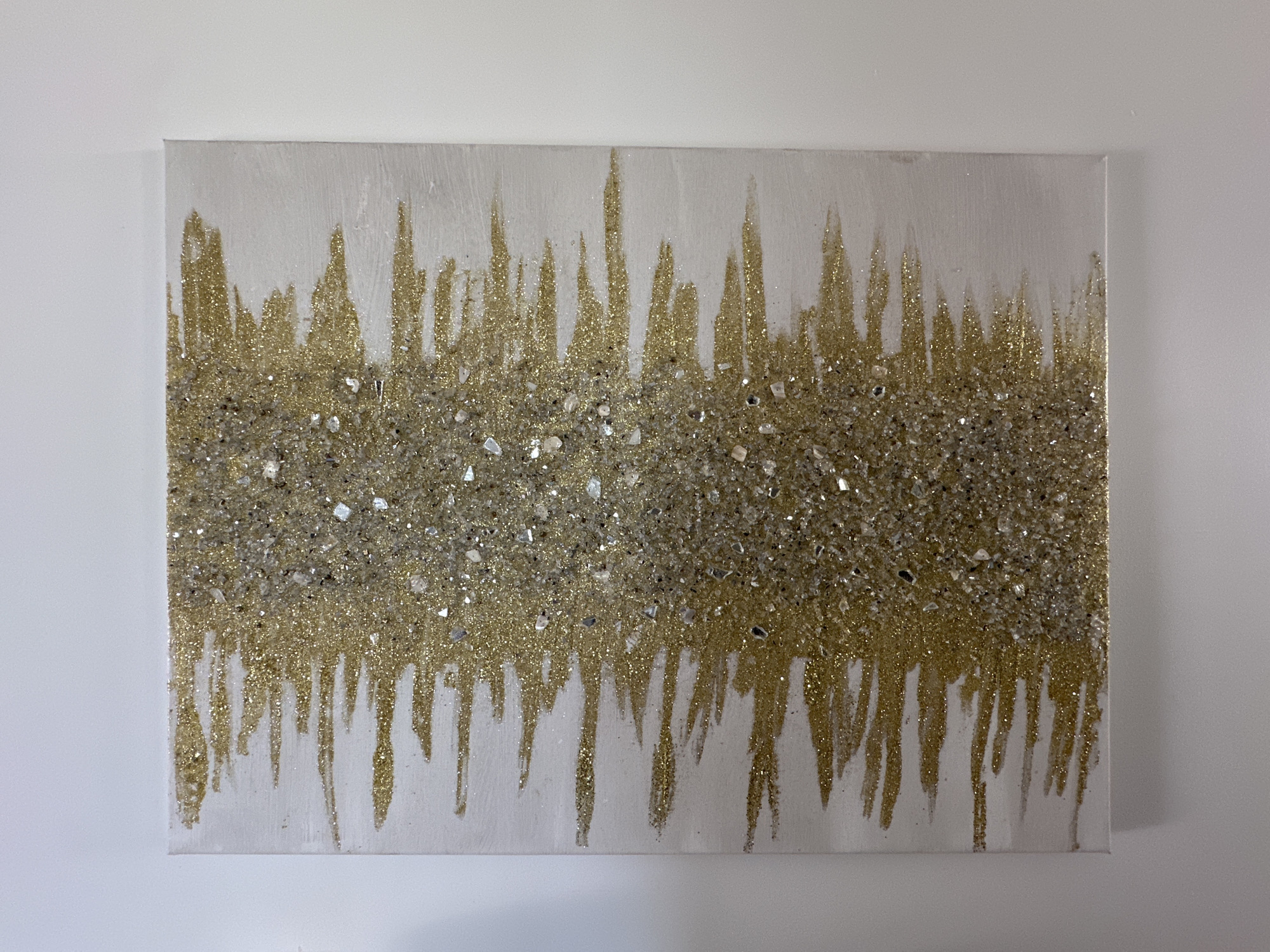 The scene is situated in the city of Marcoussis, France. A rectangular abstract artwork, horizontally oriented, is mounted centrally on a plain, smooth white wall. The artwork features a white painted background. A dense, horizontal band of fine gold glitter and irregularly shaped clear, reflective glass or crystal shards spans the approximate middle of the canvas. From this central textured band, numerous irregular, narrow vertical streaks of fine gold glitter extend upwards towards the top edge and downwards towards the bottom edge, resembling drips. The gold glitter and reflective shards provide a highly textured, sparkling surface to the artwork. The wall surrounding the canvas is devoid of other objects or markings. Even, ambient lighting illuminates the scene, highlighting the reflective qualities of the artwork's materials.