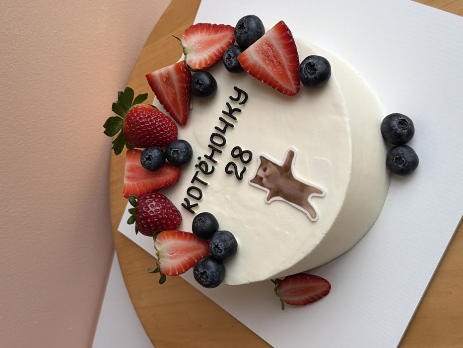 A white frosted cake is decorated with fresh strawberries and blueberries. Cut strawberry halves and whole blueberries are arranged around the top edge. In the center of the cake, black lettering spells out "Kotehochky 28", and there is a small illustration of a brown and white cat. A sliced strawberry sits on the white board beneath the cake, which is placed on a wooden surface next to a peach-colored wall.