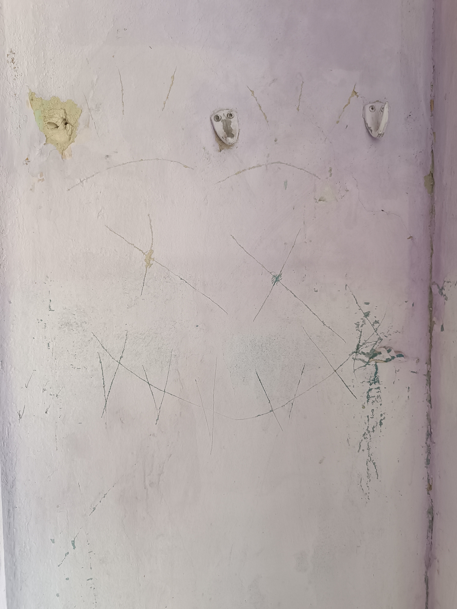 The image shows a section of a weathered, light purple-colored wall in Yelets, Russia. The wall exhibits signs of damage and age, including peeling paint and a large, irregular patch where the plaster is missing, revealing a yellowish material underneath. Several faint, scratched markings are visible across the wall's surface, some resembling abstract lines and others appearing as crude drawings. Two small, white, plastic wall fixtures, resembling stylized faces with two circular indentations for eyes, are mounted on the wall. One fixture is to the left, near the damaged plaster, and the other is to the right, further down the wall. The lighting suggests an indoor or sheltered outdoor environment. The overall impression is one of disrepair and neglect.
