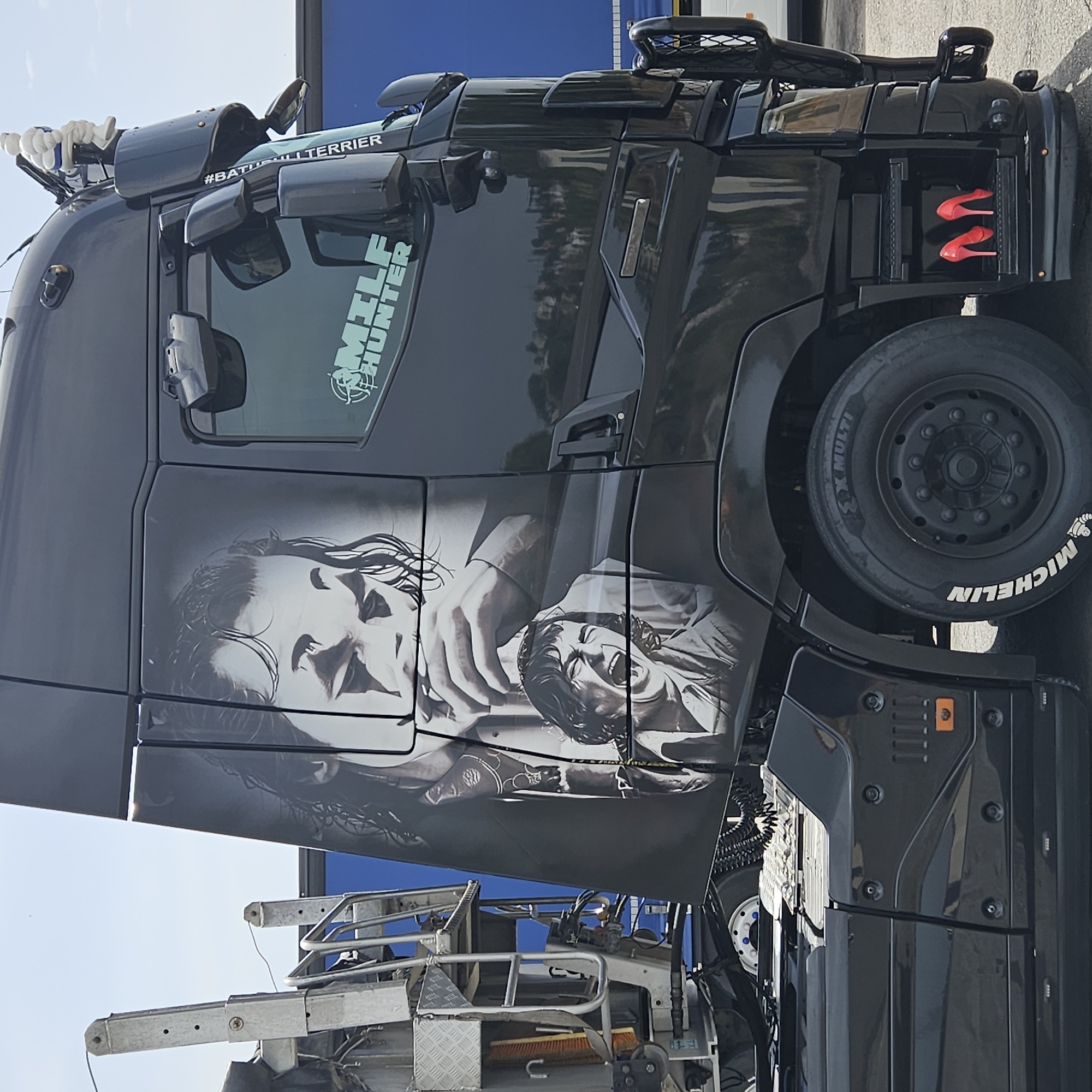 The image shows a black semi-trailer truck parked outdoors. The truck has a large graphic of a man's face, resembling the Joker, on its side. There are also smaller graphics on the truck, including the words "MILF HUNTER" and "#BULLTERRIER". A white Michelin Man figurine is perched on top of the truck. Red high-heeled shoes are visible in a compartment on the side of the truck. The background shows a clear sky and a blue trailer. The scene appears to be in a truck stop or industrial area, possibly in the city of Torrent, Spain, given the context provided.