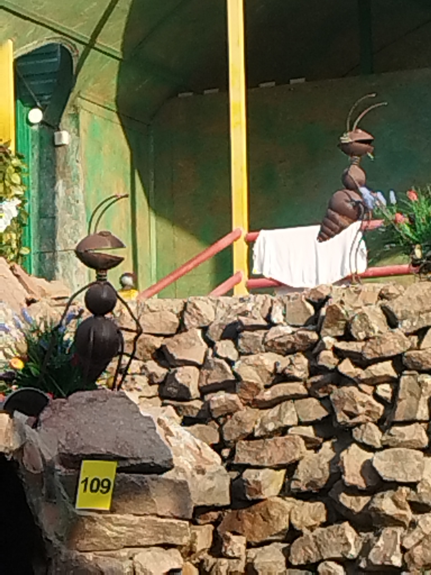 The image shows two metal sculptures of ants on a stone wall in Shymkent, Kazakhstan. The ant on the left is larger and in the foreground, with its body made of dark, rounded metal segments and its head featuring open jaws and antennae. It is positioned on a rocky surface, with a yellow sign displaying the number "109" attached to a rock in front of it. The ant on the right is smaller and further back, also made of dark metal, and appears to be resting or sitting on a red railing draped with a white cloth. Behind the ants, the environment is predominantly green, with yellow and red elements that might be part of a structure or decoration. There are also some flowering plants visible in the background. The overall scene suggests an outdoor decorative or artistic display.