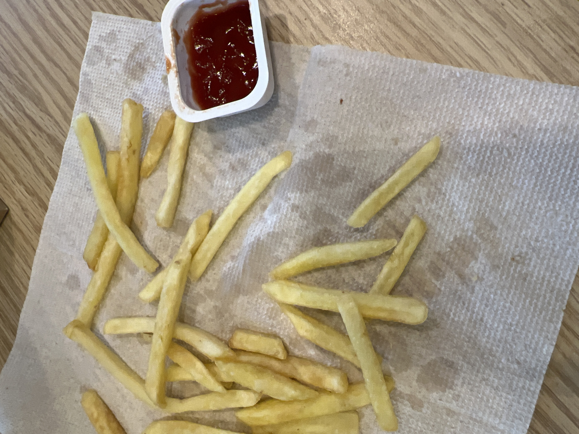 The image shows a pile of french fries scattered on a paper napkin, with a small white container of ketchup to the right. The fries are golden brown and appear to be fresh. The napkin is placed on a wooden surface, possibly a table. There are no people visible in the image. The scene is likely a casual dining setting, such as a fast-food restaurant or a picnic. There is no indication of the location in Yekaterinburg, Russia.