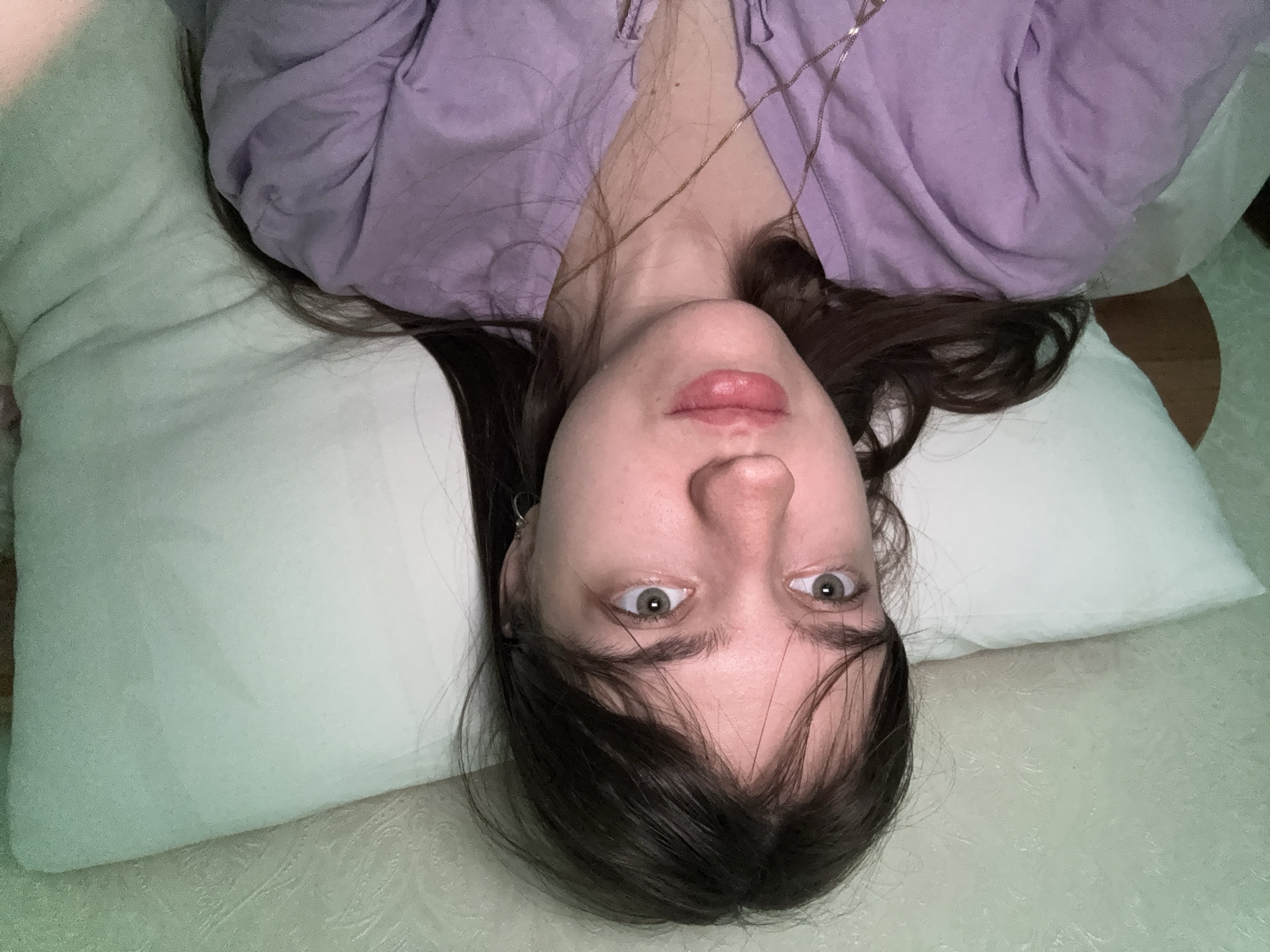 A person is lying down, facing the camera. They are wearing a light purple shirt and a necklace. Their head rests on a white pillow. The setting appears to be a bedroom. The location is unknown.