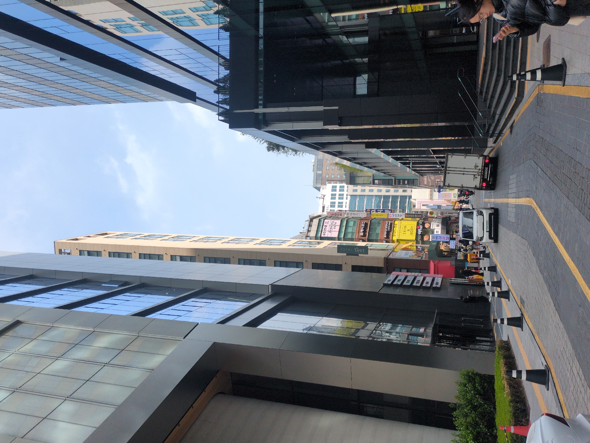 The image captures a street in Seoul, South Korea, framed by modern buildings. The left building features a reflective glass facade, while the right building is darker, with steps leading up to it. In the foreground, there are black and white striped traffic cones lining the curb. A person is visible on the right, looking at a mobile phone. The street shows light vehicular traffic, including a white delivery truck. Various commercial signs are visible on the buildings further down the street, with text in Korean. The sky is partially obscured by the buildings, showing a few clouds.