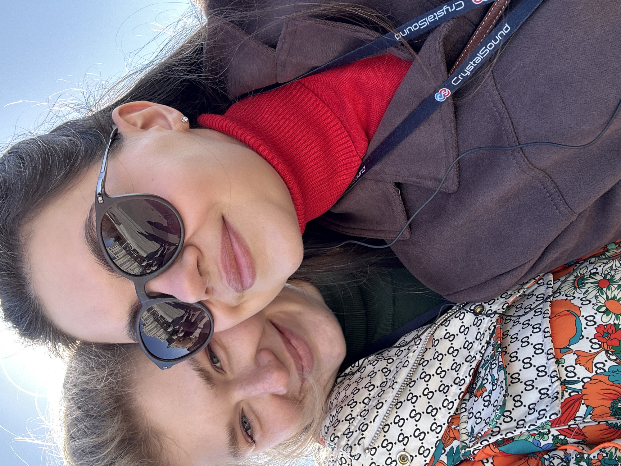 Two light-skinned women pose closely together outside in daylight. The woman at the top has dark hair, sunglasses reflecting buildings, and wears a red turtleneck under a brown jacket with a "CrystalSound" lanyard. The woman at the bottom has light hair and wears a patterned jacket with a floral and "G" logo design. The sky is visible. The reflected buildings in the sunglasses suggest a city. The location is Kazan, Russia.