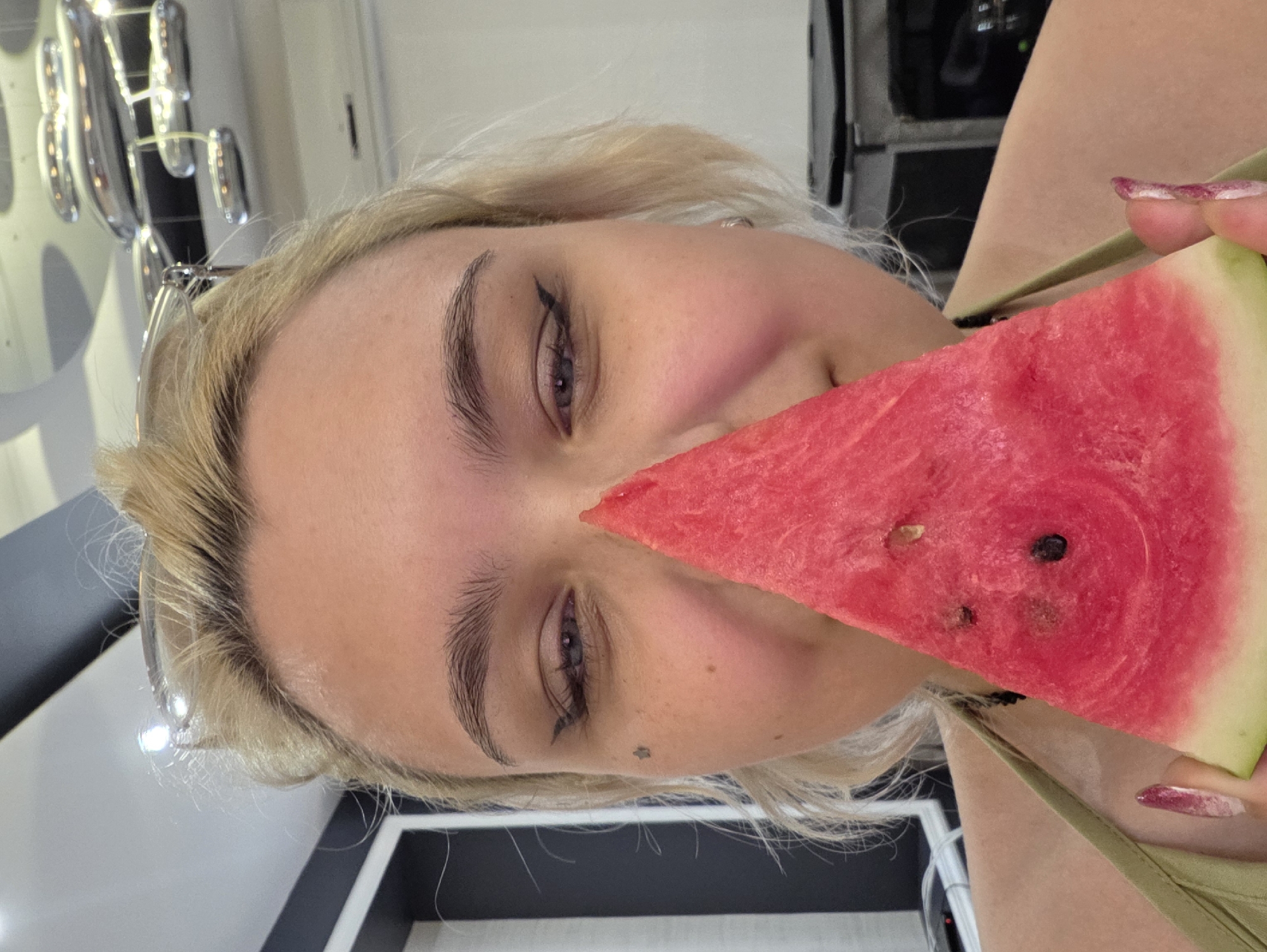 The image shows a close-up of a young woman with blonde hair and fair skin, holding a triangular slice of watermelon in front of her face, obscuring her nose and mouth. She has her eyes open, with visible eye makeup, including winged eyeliner. A small star tattoo is visible on her left cheek. She is wearing a light green top. The background is out of focus, but appears to be an indoor setting, possibly a room with modern light fixtures. There are no visible indicators of the location being within the city of Nagorny, Russia.
