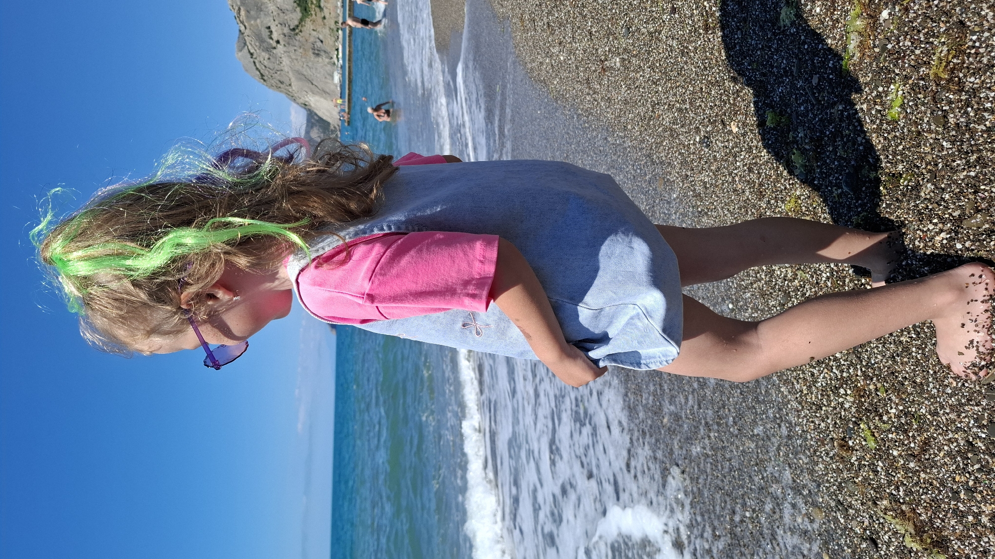 An eye-level, medium shot captures a young girl standing on a pebbled beach, facing the ocean. Her blonde hair, styled in a ponytail, is adorned with streaks of bright green and purple tinsel. She wears purple heart-shaped sunglasses, a bright pink t-shirt, and a light blue denim pinafore dress. Her left hand is tucked into the front of her dress. The sea, with small waves breaking on the shore, is visible in the background, alongside a distant rocky cliff and a pier. Several people are seen in the water and on the pier in the background. The sky is clear and blue, indicating a sunny day.