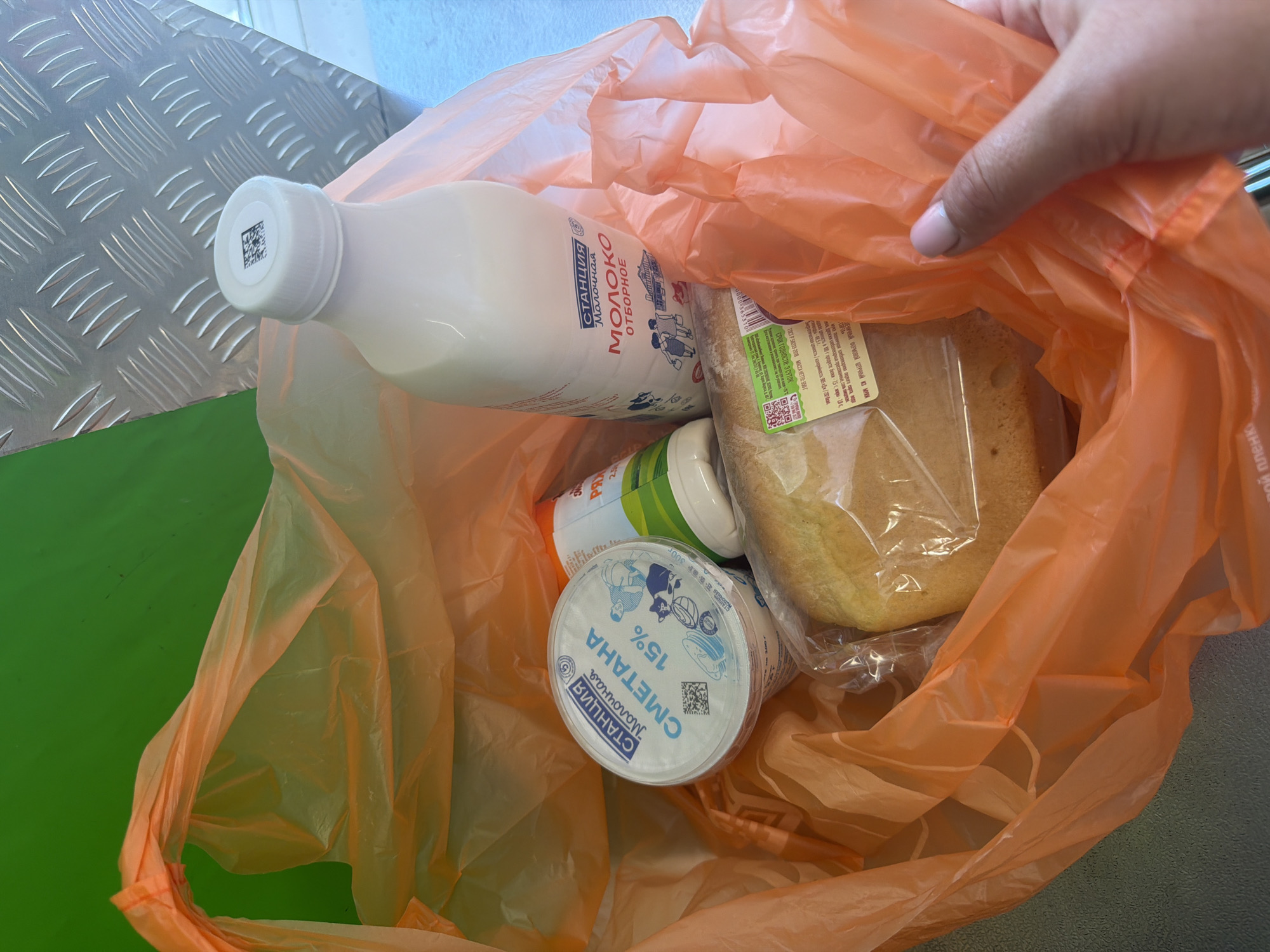 In this close-up, overhead shot, a hand is holding an orange plastic shopping bag filled with groceries. The bag's contents are partially visible: a white bottle with a QR code on its cap, a round container labeled "СМЕТАНА 15%" (Sour Cream 15%), a loaf of bread wrapped in plastic, and another cylindrical item with green and white labeling. The milk bottle has text in Russian, including "Молочная СТАНЦИЯ" (Milk Station) and "ОТБОРНОЕ МОЛОКО" (Selected Milk), suggesting dairy products. The sour cream container also bears the "Молочная СТАНЦИЯ" branding. The background includes a metallic surface with a diamond plate pattern and a green surface. The lighting suggests an indoor setting, possibly a supermarket or a home kitchen. The visible text indicates that the location is in Leningradskaya, Russia. The image captures a moment of carrying groceries, likely after a shopping trip.