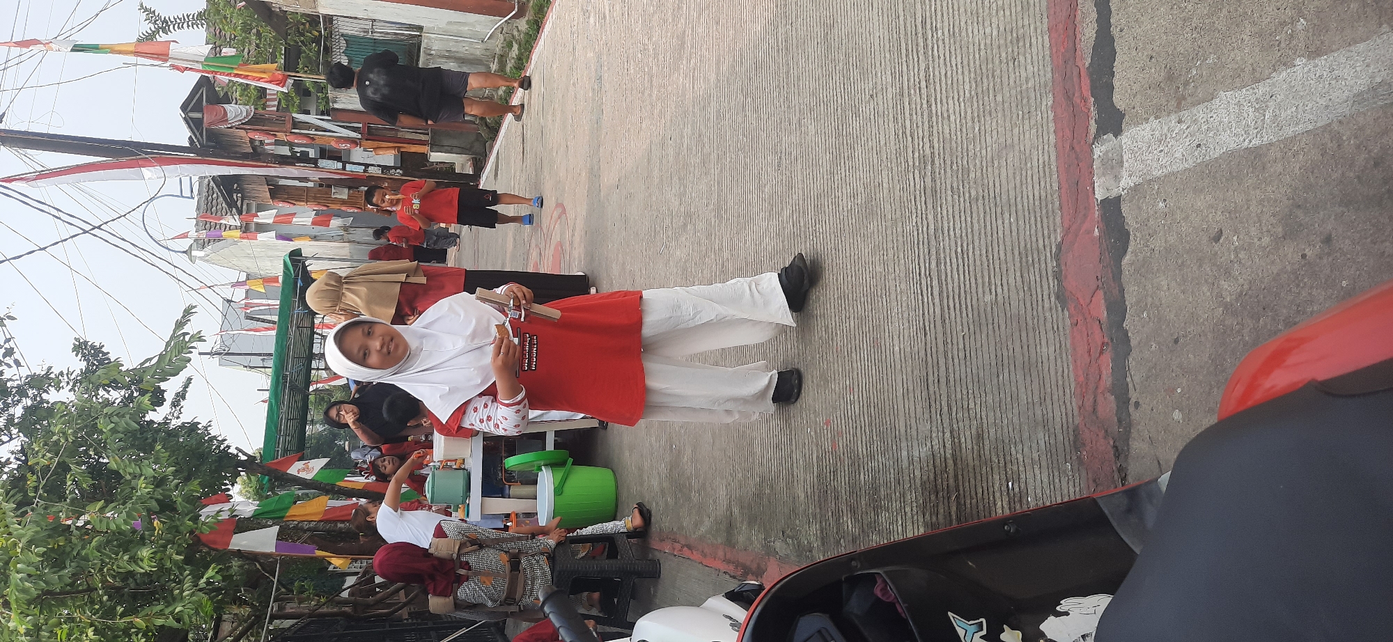 In Cisauk, Indonesia, a young girl wearing a white hijab and a red and white shirt stands on a paved street, holding a small object. She is looking directly at the camera with a slight smile. In the background, a group of people, including children and adults, are gathered. Some are wearing similar attire, suggesting a community event or celebration. Indonesian flags are visible, decorating the street and buildings, indicating a national holiday or cultural gathering. The environment appears to be a residential area with buildings and trees lining the street. A scooter is parked in the foreground, partially obscuring the view of the street and the people. The overall scene depicts a lively and festive atmosphere.