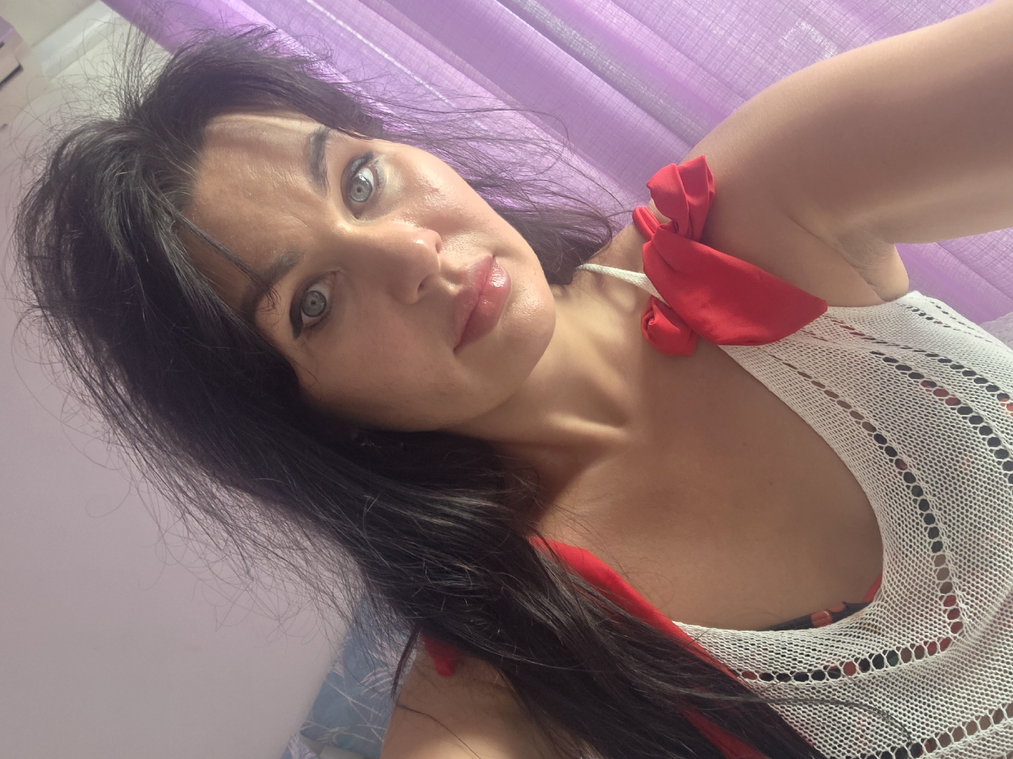 A woman with long, dark hair and blue eyes, wearing a white mesh top with red ribbon accents on the shoulders, is pictured in a selfie. She has light makeup on, including eyeliner and lip gloss. The background consists of a light-colored wall and a purple, sheer curtain. The setting is an indoor space, possibly a bedroom, with no specific identifying markers of Sochi, Russia visible in the image.