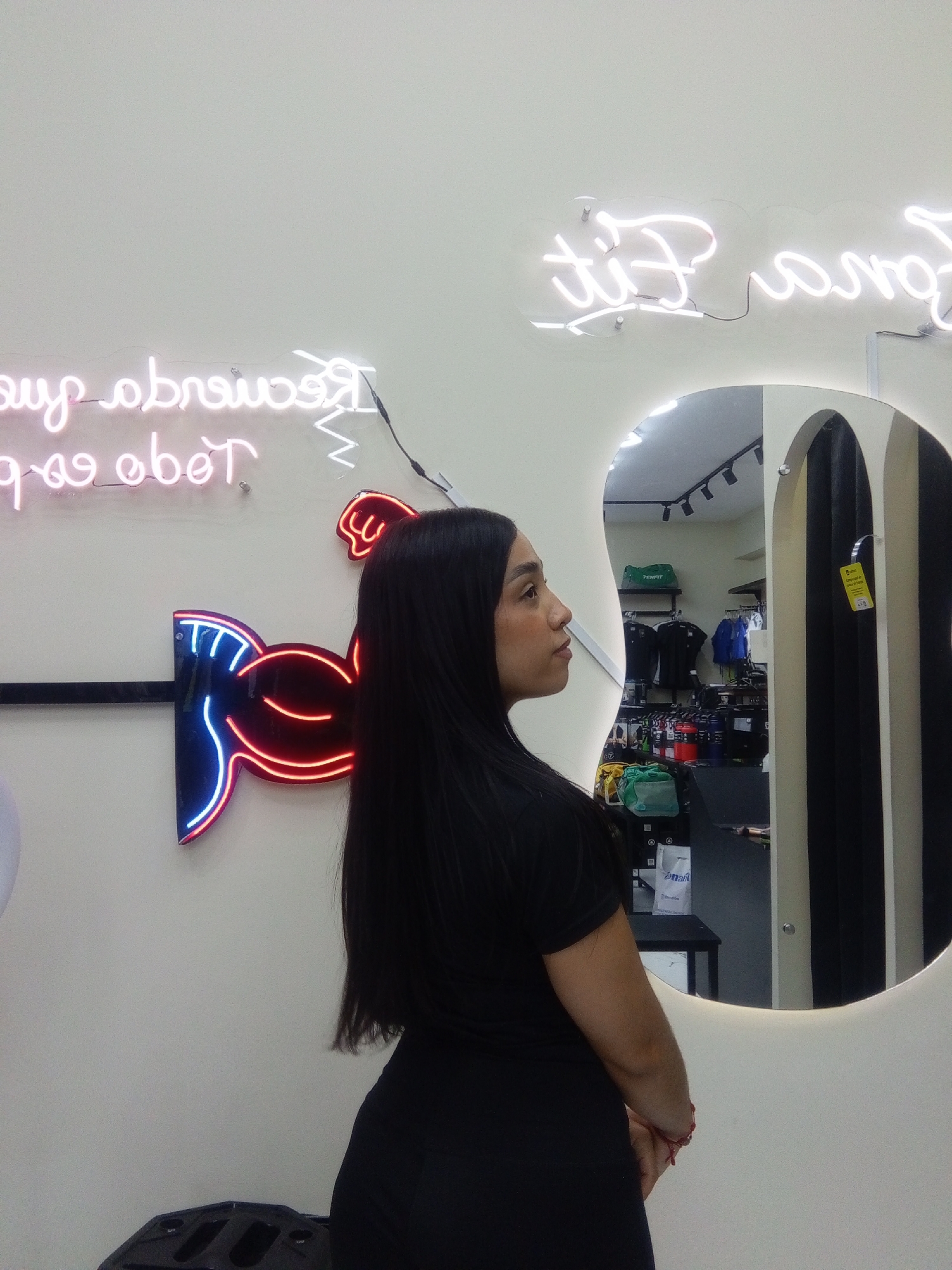 The image shows a woman standing indoors, with her back partially turned to the camera, looking towards her right. She has long, dark, straight hair and is dressed in a black t-shirt and dark bottoms. A red bracelet is visible on her left wrist.
She is positioned in front of a light-colored wall featuring multiple neon signs. On the upper left, a pink neon sign displays reversed text that appears to read "Reactivando tus pasos todo". Above a mirror to the right, a white neon sign also shows reversed text, "Para Fit". Below the pink text, there is a prominent neon graphic in the shape of a flexing bicep or arm, glowing in red and outlined in blue.
To the woman's right, a large, organically shaped mirror reflects the interior of what appears to be a fitness or athletic wear store. The reflection shows clothing racks with black and blue t-shirts, green gym bags, and shelves stocked with red and black canisters or bottles, likely supplements. Overhead track lighting is also visible in the reflection. A yellow tag hangs from an item within the reflection, though its text is illegible.
The setting is a brightly lit indoor space, strongly suggesting a retail establishment focused on fitness products, likely in Valle de La Pascua, Venezuela. The woman's expression is neutral and focused.