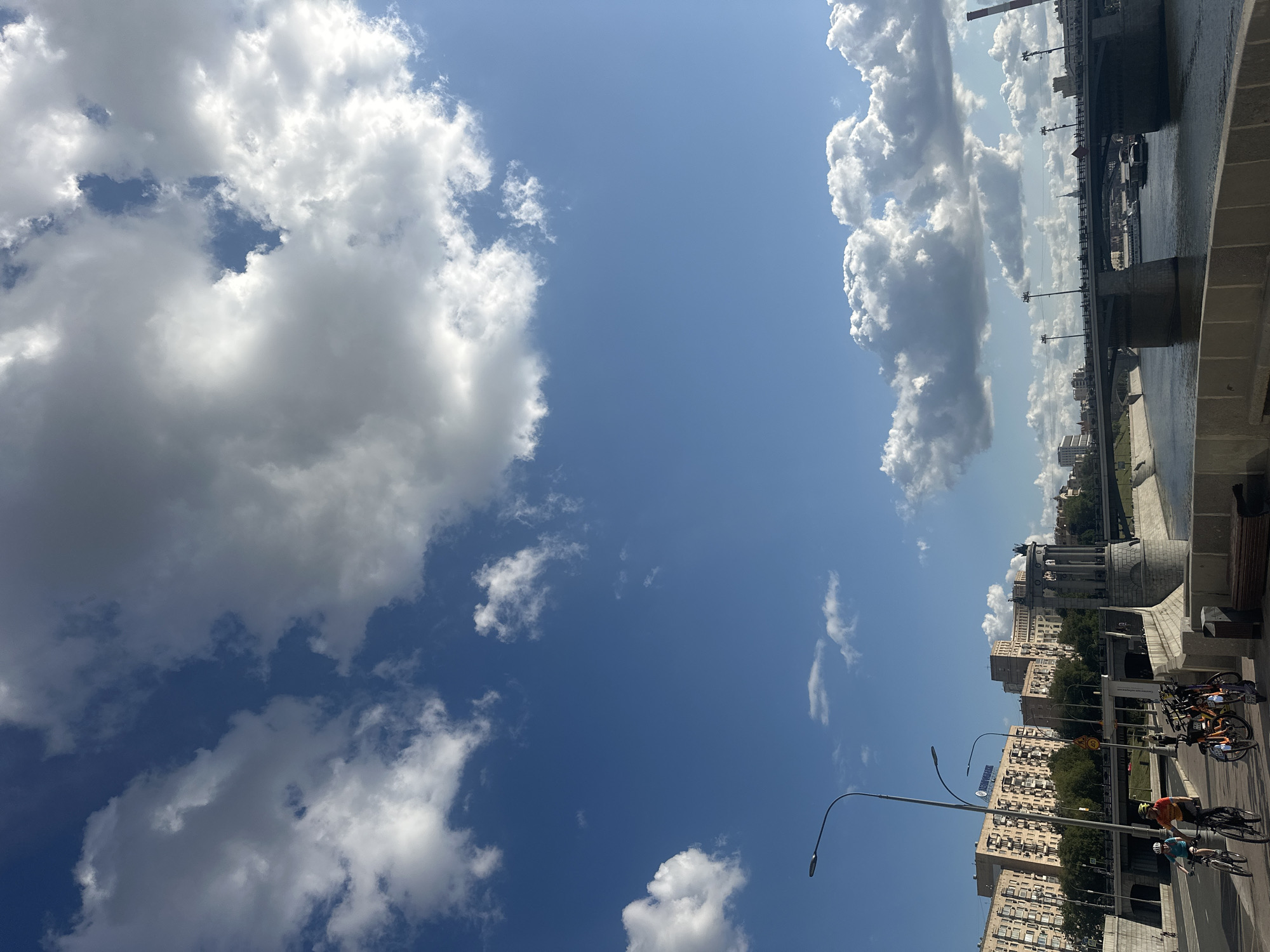 This image depicts a sunny day in Moscow, Russia, with a clear blue sky dotted with fluffy white clouds. The scene is dominated by a wide river, likely the Moskva River, flowing through the city. A multi-lane bridge spans the river in the mid-ground, with traffic and pedestrians visible on its deck. On the left bank of the river, a paved embankment is visible, lined with trees and a sidewalk where several people are cycling. In the background, apartment buildings and other city structures are present. A prominent circular, colonnaded structure stands on the embankment near the bridge. The overall impression is of a lively urban landscape on a pleasant day.