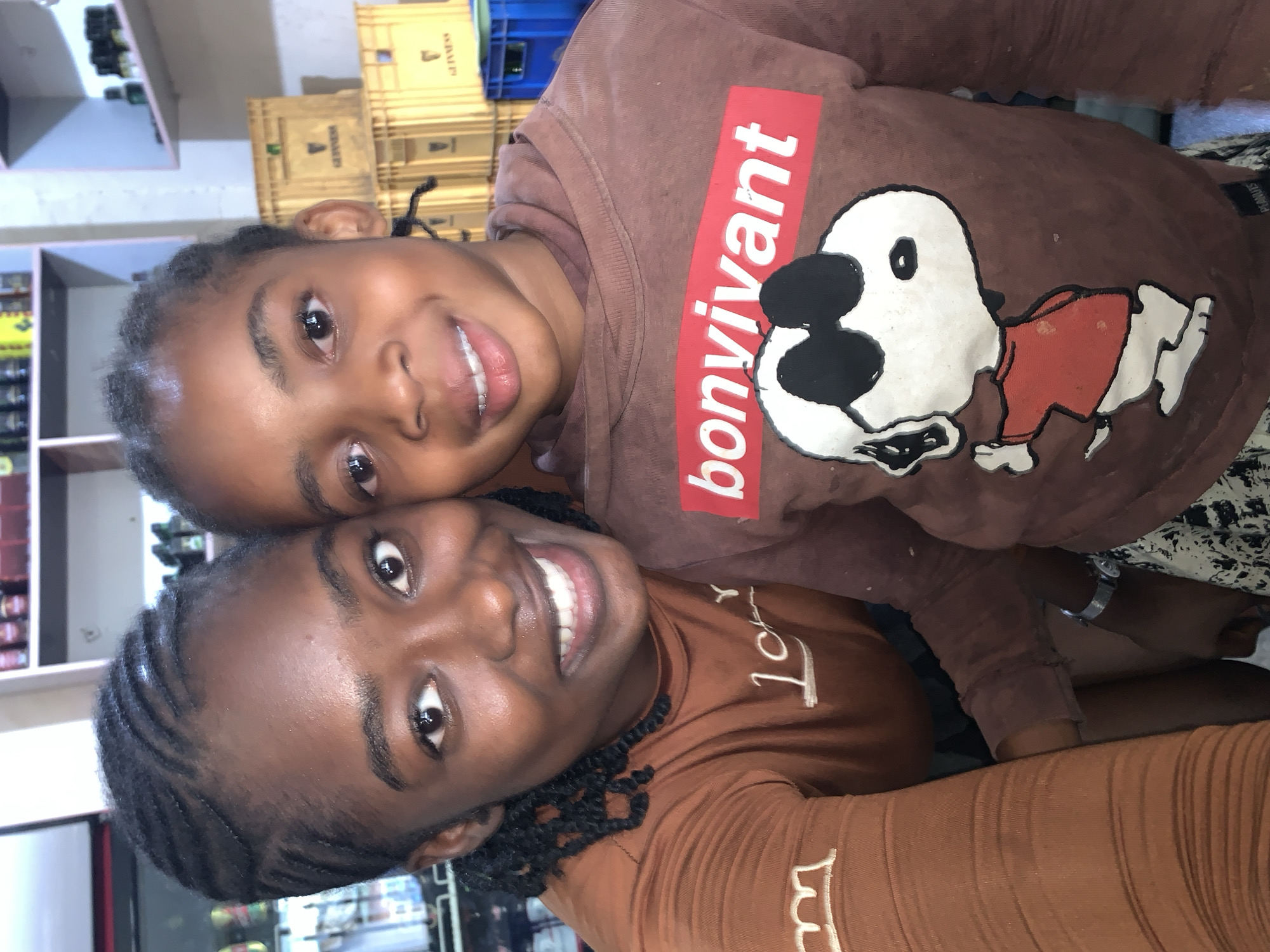 Two individuals, an adult female and a female child, are depicted in a close-up, front-facing selfie portrait, captured indoors in what appears to be a commercial or storage area in New Stadium, Nigeria. The adult female, positioned on the left, exhibits dark skin, dark eyes, and a broad smile with visible teeth. Her hair is styled in cornrows with intricate patterns on the sides of her head, and some braids are visible around her neck. She wears a rust-brown long-sleeved top featuring white embroidered details on the left sleeve and chest. Her left arm is partially visible, extending towards the child. The child, positioned on the right and slightly in front of the adult, also has dark skin, dark eyes, and a smile with visible teeth. Her hair is styled in small braids pulled back from her face. She wears a dark brown hooded sweatshirt with a prominent red rectangular graphic on the chest displaying the white text "bonvivant." Below this, a white cartoon graphic of Snoopy, wearing black sunglasses and a red shirt, is visible. A silver-toned wrist watch is visible on a hand near the child's lower right side. The child's lower body is partially obscured by a black and white patterned garment. The background features light-colored shelves displaying numerous bottles and containers. To the right of the child, several stacked yellowish-brown crates, each bearing the "GUINNESS" logo, are visible, alongside a partially visible blue plastic crate. The scene is evenly lit, indicating an indoor environment.