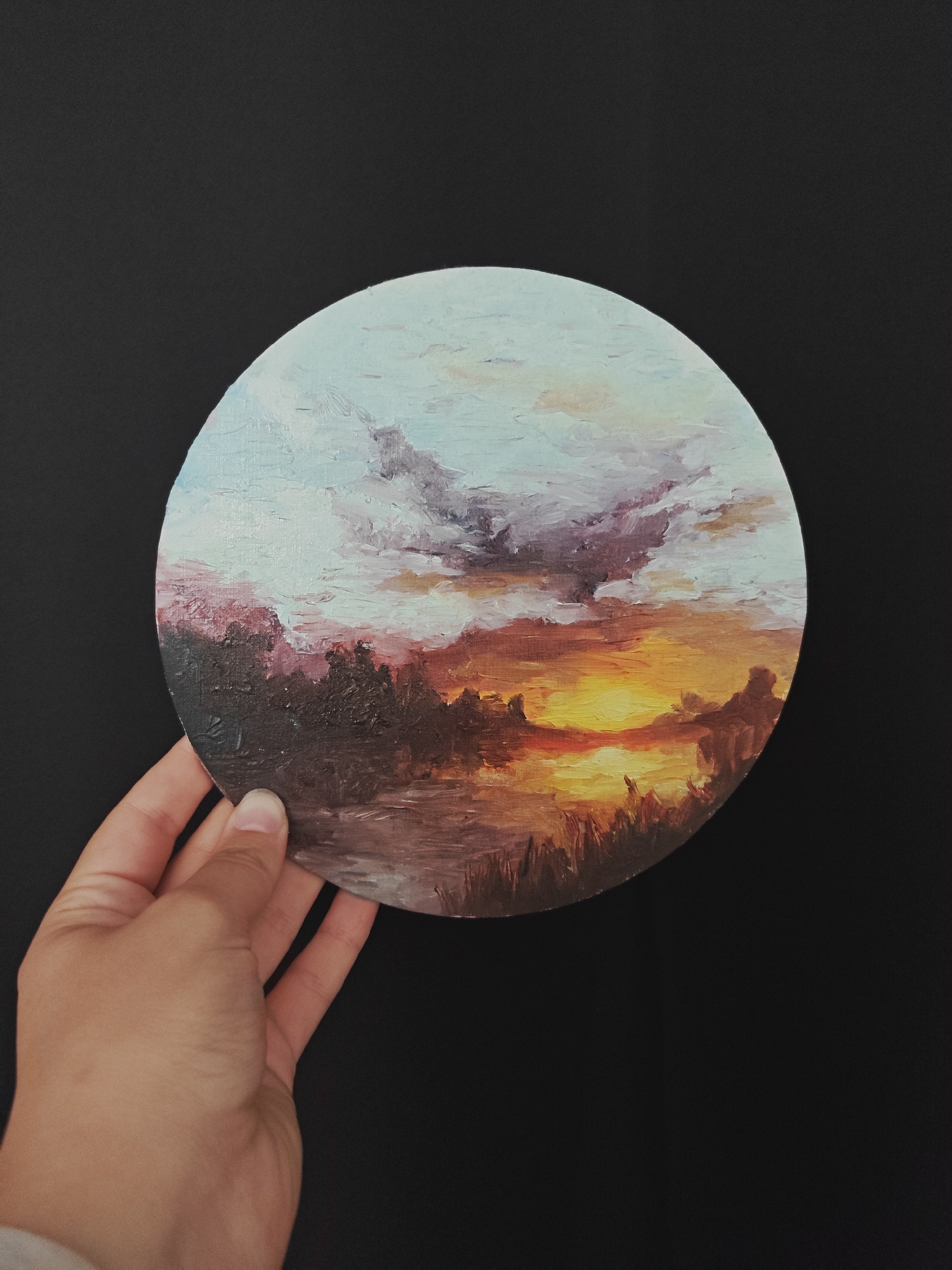 A hand holds a circular, textured oil painting of a vibrant sunset over a landscape. The painting depicts a fiery orange and yellow sun sinking below a dark, silhouetted treeline. The sky above is a blend of soft blues, purples, and pinks, with dramatic clouds. The foreground of the painting shows a body of water reflecting the sunset colors, with hints of vegetation along its edge. The painting is presented against a plain black background. There is no visible connection to the city of Koledino, Russia.