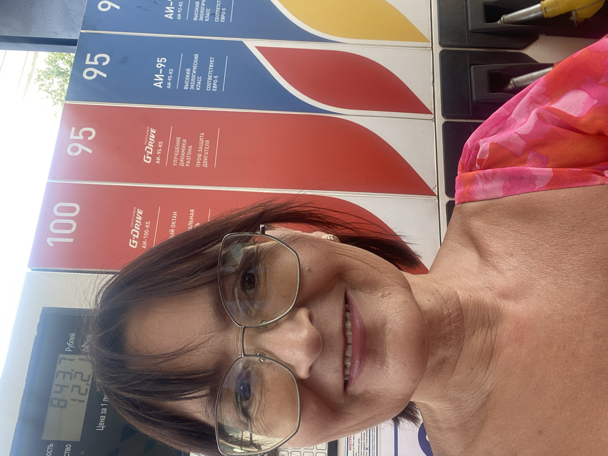 A woman is taking a selfie at a gas station. She is wearing glasses and a pink and red floral top. Behind her, there are gas pumps with prices displayed. The price displayed is 84.37 rubles per liter, and the amount dispensed is 12.2 liters. The gas station is located in Novokuznetsk, Russia.
