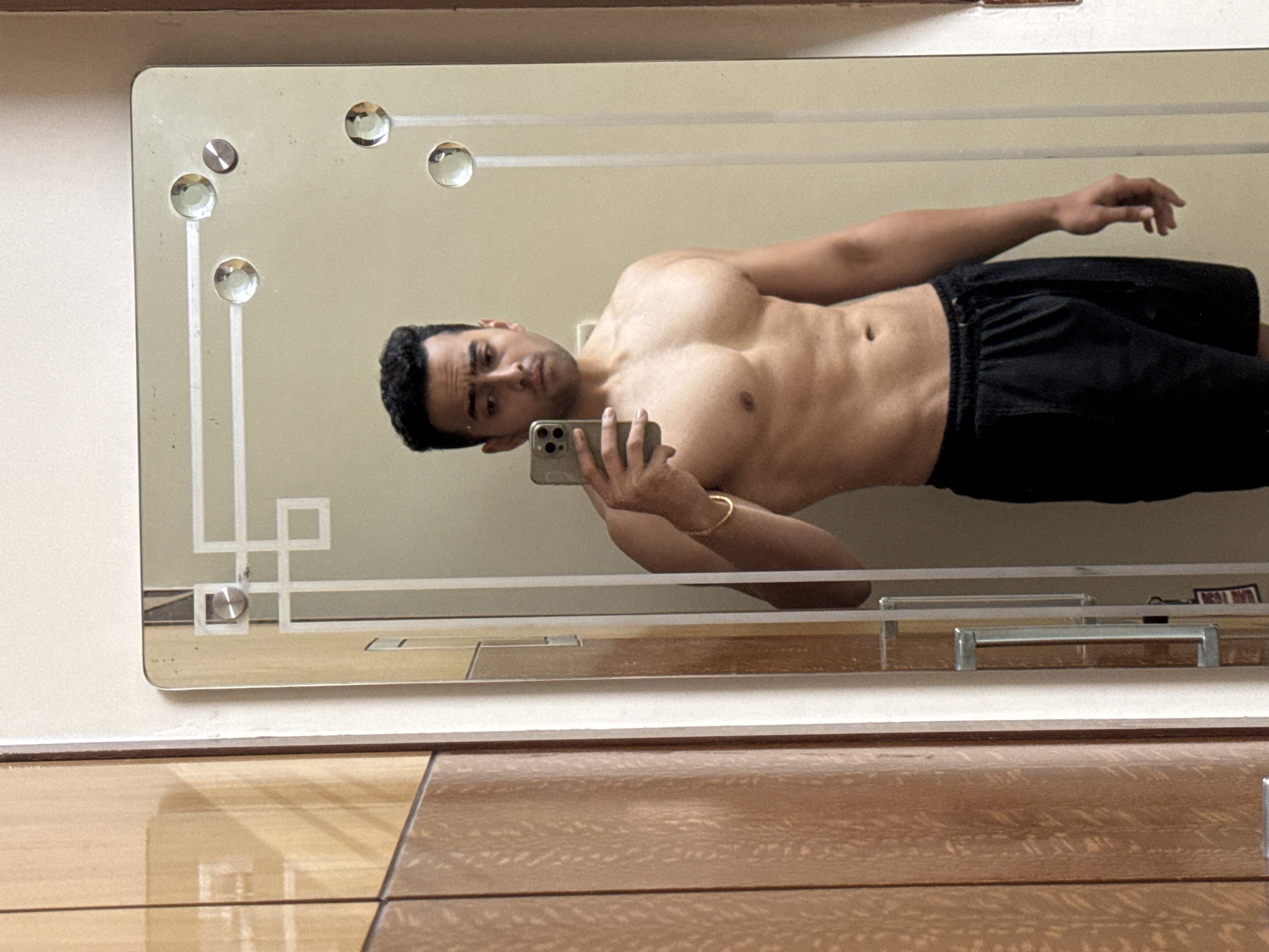 A shirtless man with dark hair and a serious expression is taking a selfie in a mirror. He is wearing black shorts and a gold bracelet on his left wrist. The mirror is mounted on a light-colored wall, and the reflection reveals a portion of a wooden dresser with drawers. The mirror has decorative elements, including small clear spheres and geometric patterns. The location could be Kolkata, India, based on the possible appearance of furnishings that can be found there.
