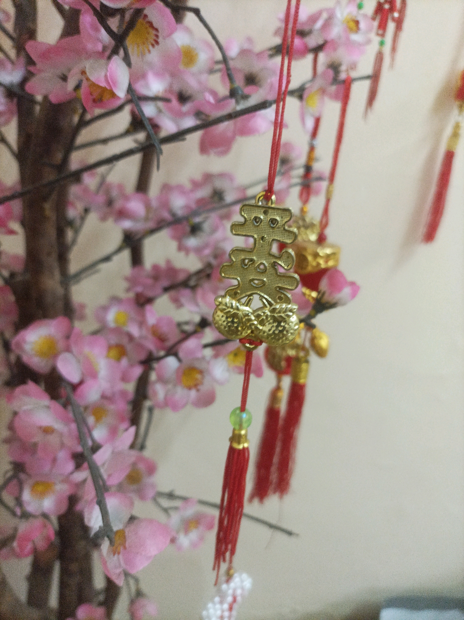In Bekasi, Indonesia, a close-up shot captures a decorative artificial plum blossom branch adorned with traditional Chinese New Year charms. The branch features numerous delicate pink blossoms with yellow centers, interspersed with brown branches. Hanging from red strings are golden charms, including one intricately designed character symbol and two clusters of golden fruit. Red tassels and small green beads are also visible, adding to the festive ornamentation. The background is a plain, light-colored wall. The overall scene suggests a cultural decoration, likely for the Lunar New Year celebration.