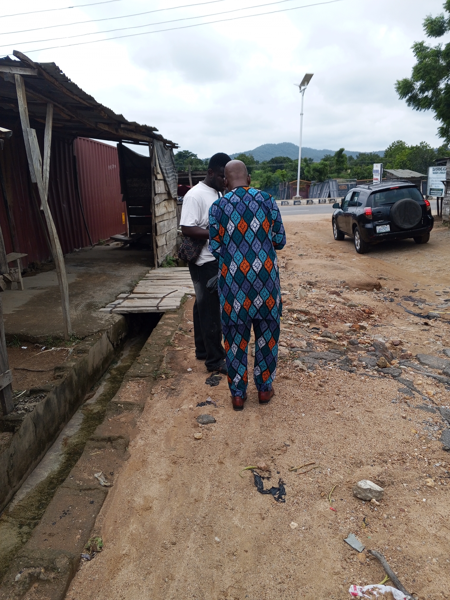 In Zango-ndaji, Nigeria, two men are engaged in conversation on a dusty, unpaved road next to a rudimentary structure. The man on the left wears a white t-shirt and dark trousers, with a bag slung over his shoulder. The man on the right is dressed in a vibrant, patterned dashiki and trousers, with brown shoes. They stand facing each other, their interaction suggesting a discussion.
Behind them, the structure appears to be a makeshift shelter constructed from corrugated metal and wooden planks, with a partially exposed red shipping container visible within. A drainage channel runs alongside the road, covered in places by wooden planks. In the background, a black SUV is parked on the side of the road, and a tall street light stands prominently. Beyond the immediate area, a line of trees and hills form the backdrop under a cloudy sky. The ground is covered with loose dirt, gravel, and scattered debris, indicative of an unmaintained urban or semi-urban environment.