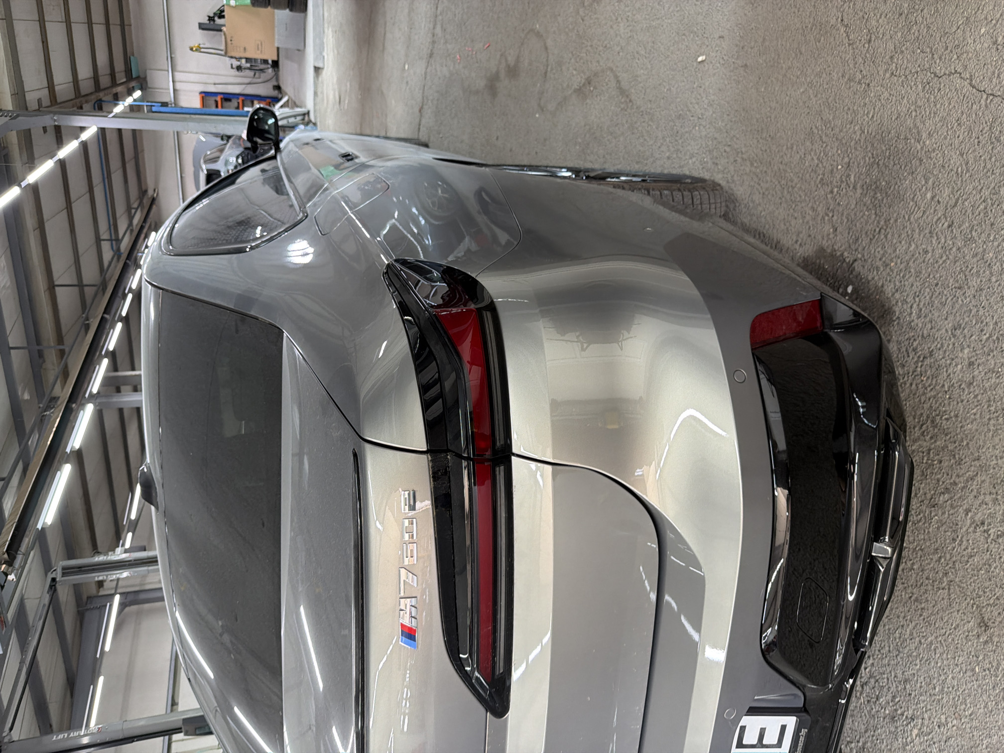 The image captures the rear three-quarter view of a sleek, grey BMW M760e, positioned indoors within a well-lit garage or workshop. The car's polished surface reflects numerous bright, elongated fluorescent lights from the ceiling, highlighting its smooth contours and pristine condition.
On the trunk lid, the "M760e" badge is prominently displayed, featuring the distinct BMW M colors of blue, purple, and red stripes. The modern, L-shaped LED taillight stretches horizontally across the rear and then turns vertically, providing a striking design element. The rear bumper incorporates both the car's grey body color and contrasting black trim elements. Small circular parking sensors are visible on the bumper. A partially visible white sticker or license plate frame on the lower right of the bumper shows the letters "WE," suggesting a possible local affiliation such as "WERA.BG".
The setting is an industrial or commercial space, likely a car service center, detailing shop, or showroom. A grey concrete floor is visible around the vehicle. Overhead, numerous rows of fluorescent strip lights illuminate the area brightly. Metal support beams and structures, possibly components of car lifts or the building's infrastructure, are visible in the background. The car is stationary, and no people are visible in the frame. The overall impression is one of a high-performance vehicle in a professional care environment, consistent with a location like Burgas, Bulgaria.