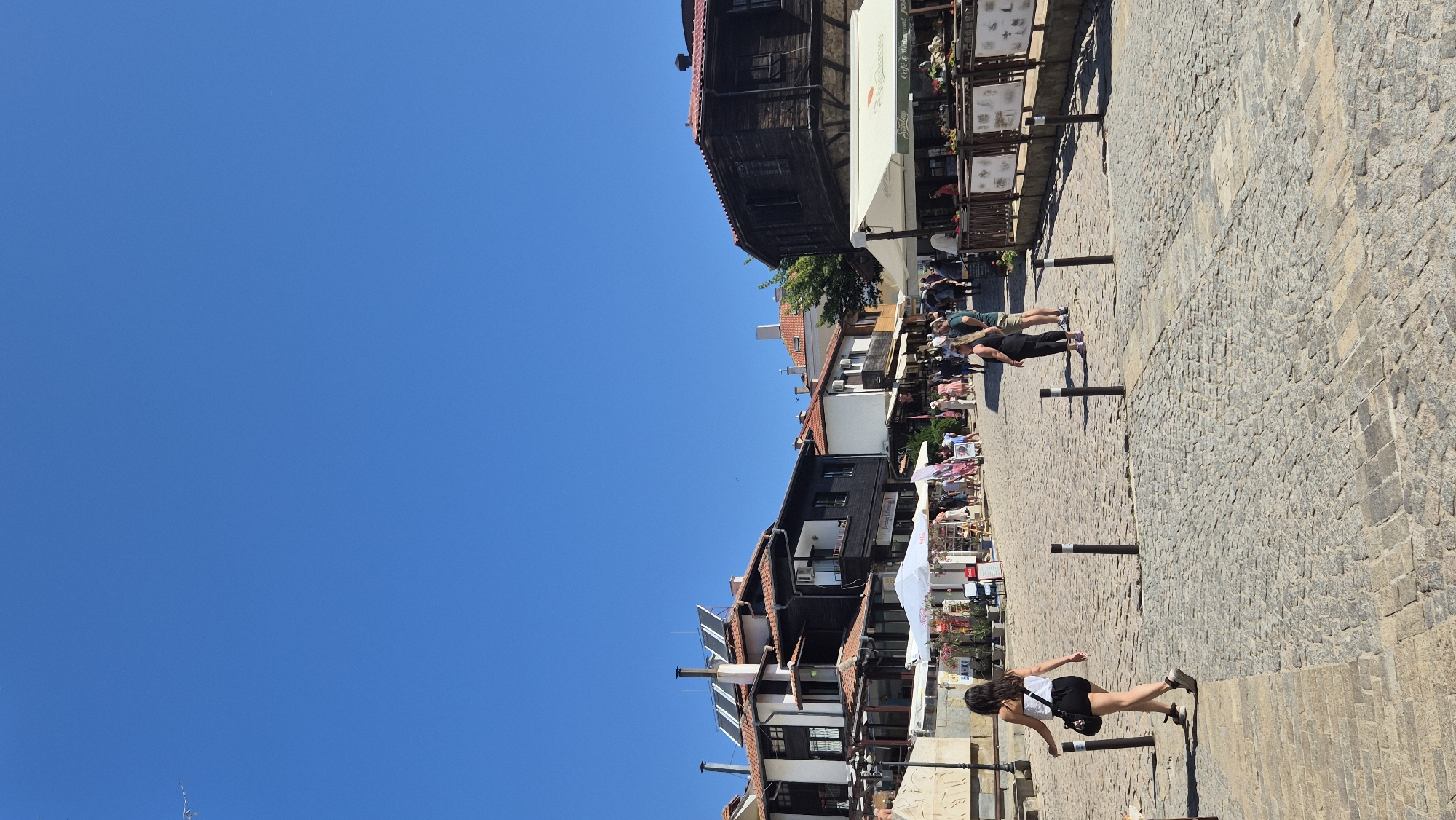 A vertical, eye-level shot depicts a bright, sunny day in the historic Old Town of Sozopol, Bulgaria, featuring a cobblestone street lined with traditional wooden and stone buildings. The sky is a clear, vibrant blue, with a faint silhouette of a bird in the upper left quadrant. In the foreground, a woman with dark hair, wearing a white tank top and black shorts, walks away from the camera, holding a dark bag over her shoulder and a black pole in her right hand. She is on the cobblestone street, marked by bollards. Several other people are visible in the middle ground, walking and browsing near outdoor stalls and cafes. One cafe, with a white awning, displays signage that reads "Cafe & Restaurant." The buildings exhibit characteristic wooden architecture, some with red tile roofs and visible solar panels on one structure. The street is paved with rough, gray cobblestones, with some sections appearing lighter. The overall scene conveys a bustling atmosphere of a popular tourist destination on a warm day.