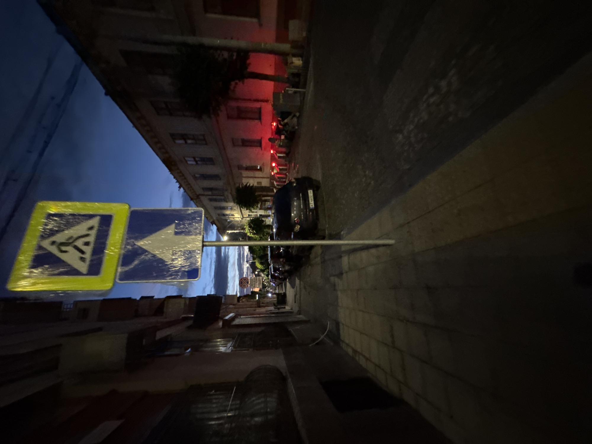 The image depicts a street scene in Chernivtsi, Ukraine, at dusk. The perspective is from a sidewalk looking down a street lined with buildings. In the foreground, a pedestrian crossing sign and a directional arrow sign are mounted on a pole. The sidewalk is paved with rectangular stones. On the right side of the street, a row of parked cars is visible, and further down, building facades with illuminated windows suggest residential or commercial use. Red lights are visible along the buildings. The sky is a deep blue with some clouds. The overall atmosphere is calm and quiet, typical of a city street in the early evening.