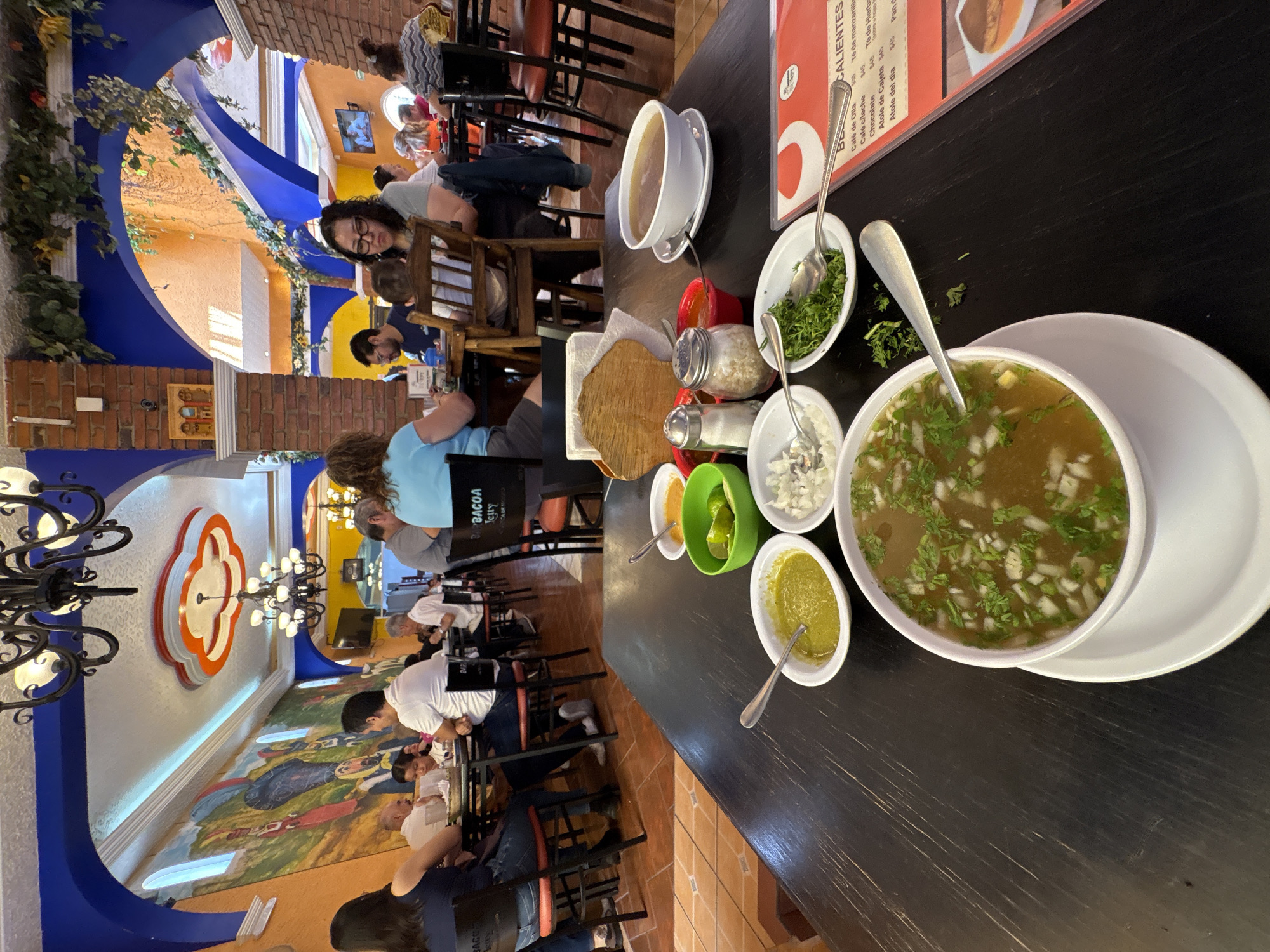 The image captures the interior of a restaurant, presumably in Ocotepec, Mexico, identifiable by its architectural style and the menu displaying "Atole de Cajeta". Several patrons are seated at tables, engaged in dining and conversation. Dishes of soup, tortillas, and various condiments are arrayed on a dark tabletop. The restaurant's décor includes a mural, ornate chandeliers, and distinctive blue and orange architectural accents.