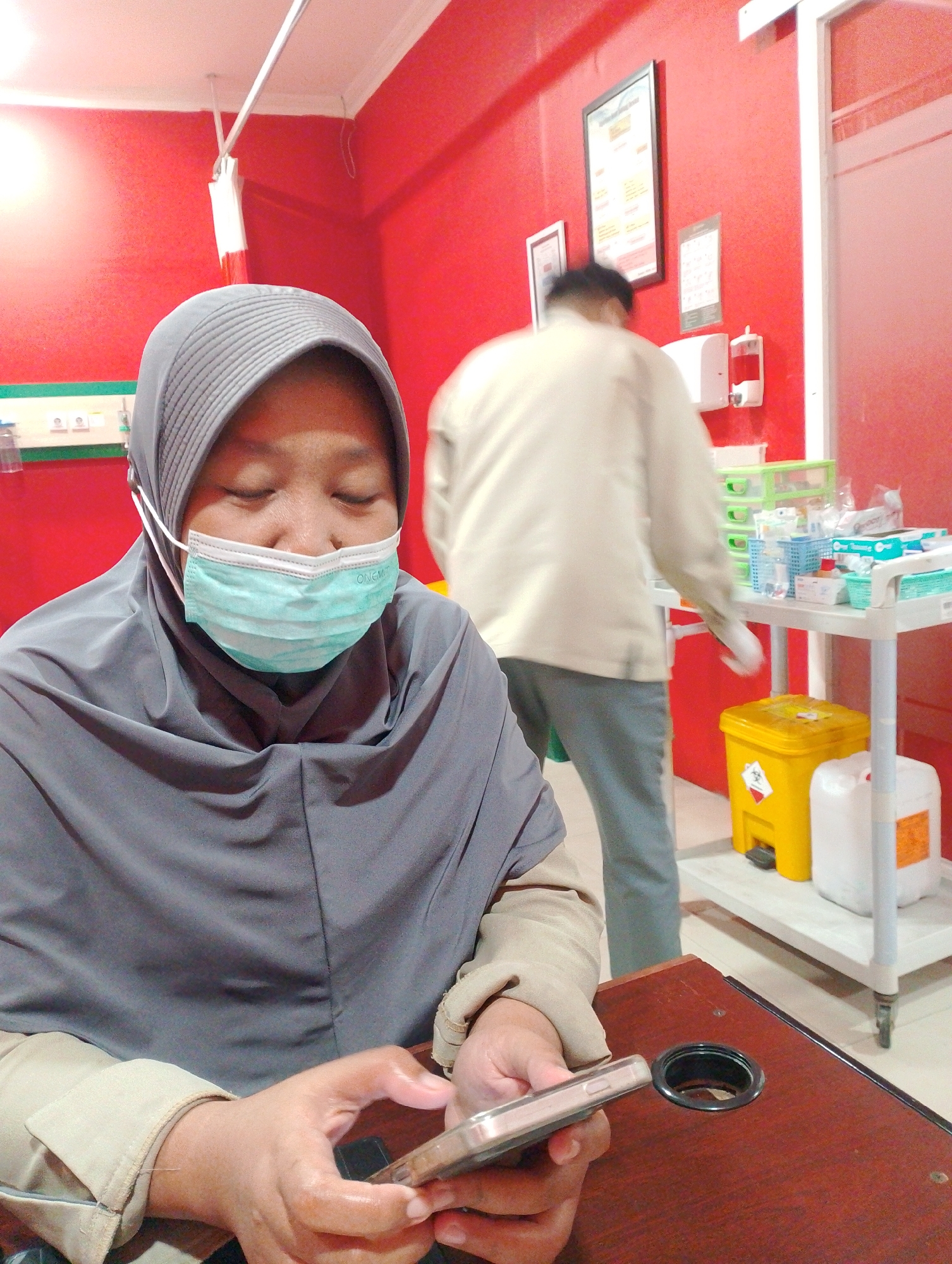 A person wearing a grey hijab and a light blue surgical mask is seated, holding a smartphone. They are wearing a tan jacket over a grey garment. In the background, another person in a light-colored shirt and grey pants is standing near a medical cart filled with supplies. The room has red walls and a white ceiling with visible lights. Medical equipment and containers are present, including a yellow biohazard bin and a white container with an orange label. A framed picture and a calendar are hanging on the wall. The overall environment suggests a medical facility, possibly a clinic or hospital in Sidoarjo, Indonesia.