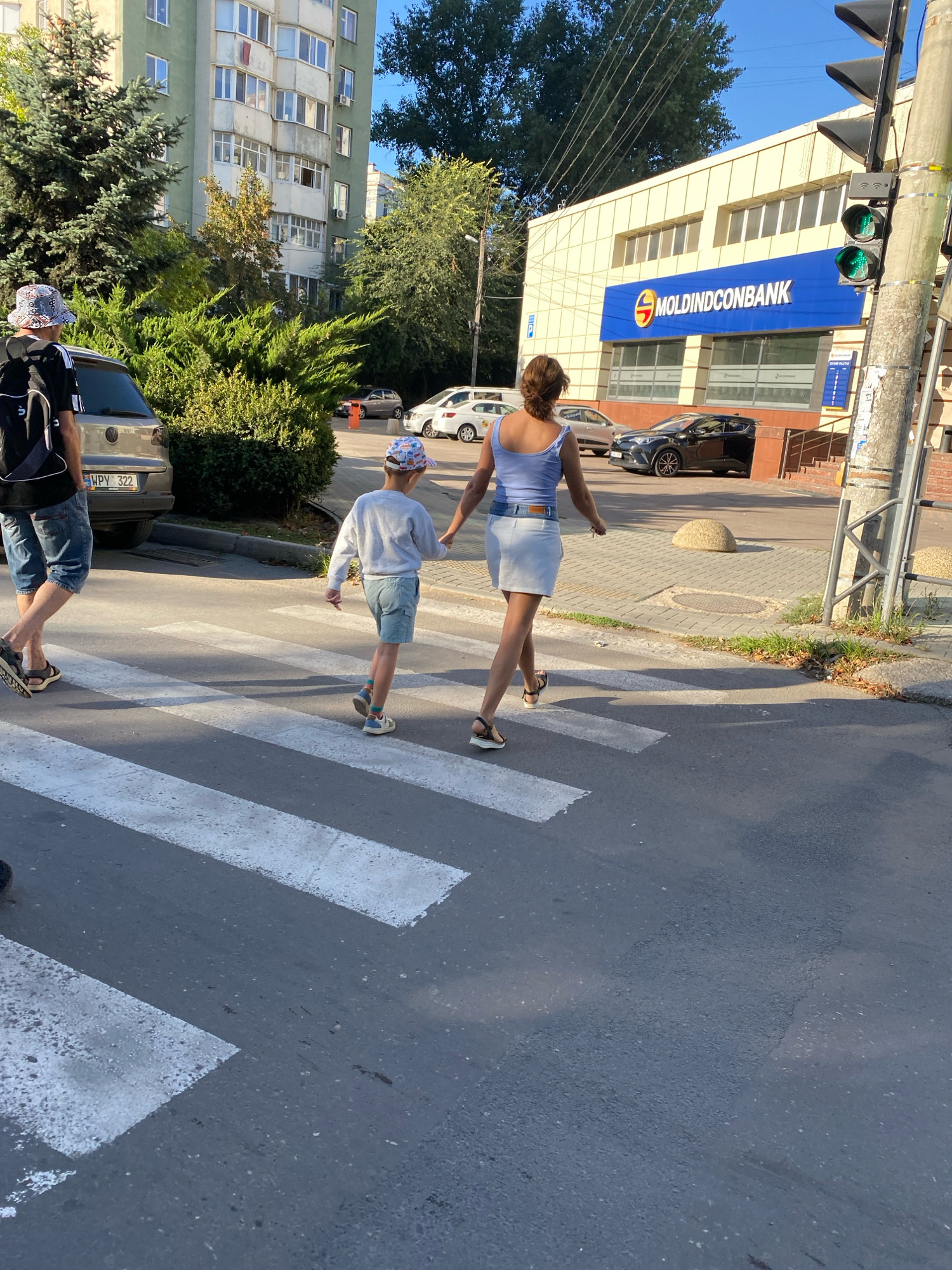 In Chișinău, Moldova, a pedestrian crossing is occupied by a woman and a young boy walking hand-in-hand. The woman, wearing a light blue tank top and a white skirt, is walking away from the viewer. The boy, dressed in a light gray long-sleeved shirt and light blue shorts, wears a patterned cap and sneakers. To the left, partially visible, is a man with a backpack, wearing a patterned hat and denim shorts. A silver Volkswagen car is parked to the left of the crossing. In the background, a building with the "MOLDINDCONBANK" logo is visible, next to a traffic light showing a green signal. The scene is illuminated by sunlight, casting shadows across the asphalt.