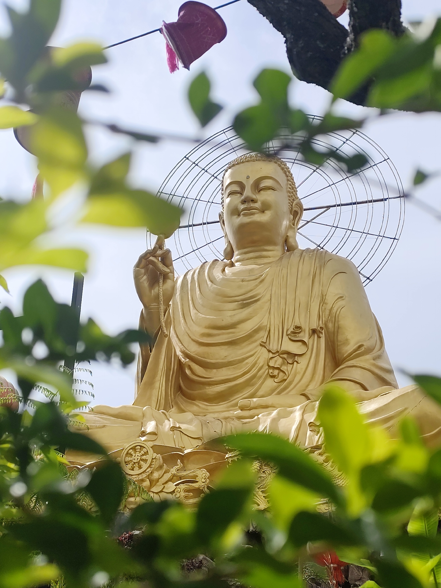 A low-angle medium shot shows a golden Buddha statue in the city of Lâm Viên - Đà Lạt, Vietnam. The statue is in a meditative seated position with a large wire halo surrounding the head. It is partially obscured by green foliage in the foreground. A string of what appears to be red and pink flower pots hangs from a wire above the statue. A tree branch is visible in the upper right corner of the image. The sky is a pale blue.