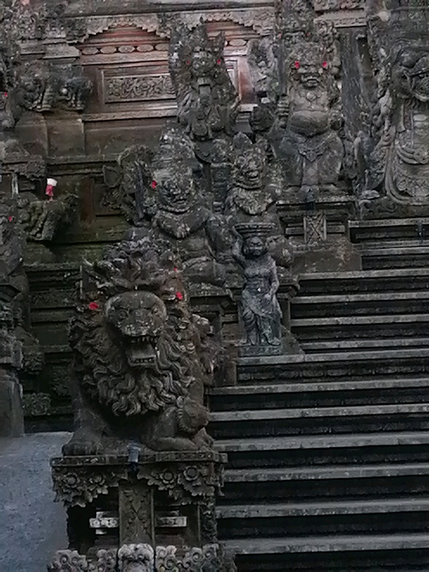 This is a close-up, low-angle, medium shot of a stone staircase adorned with intricate carvings and statues, likely within a temple complex in the Ubud District, Indonesia. The focal point is a detailed stone lion statue positioned on the left side of the frame, at the base of the stairs. The lion has an open mouth, showing teeth, and a prominent mane. Red accents are visible in the eyes of the lion and several other statues lining the stairs and the structure behind them. To the right of the lion and further up the stairs, a small, carved human-like figure stands, holding an object in its raised right hand. The stairs are made of rough, dark stone and are quite wide. The background reveals a densely carved stone facade with multiple figures and architectural elements, suggesting a significant cultural or religious site. The overall impression is one of ancient artistry and spiritual grandeur.