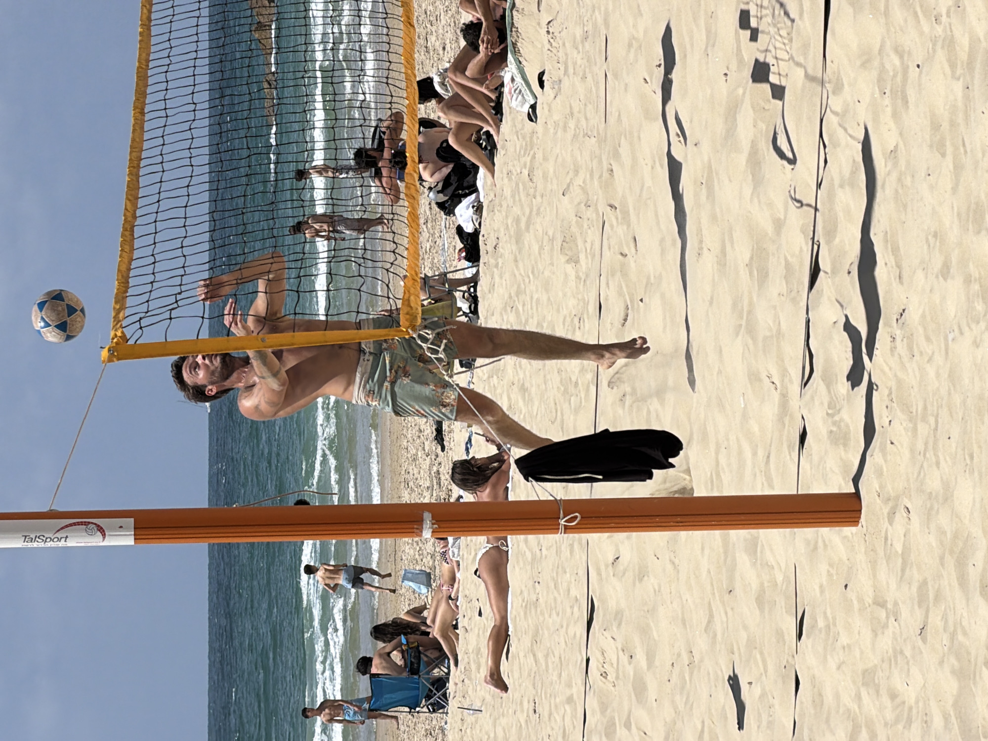 The image depicts a beach volleyball scene, likely at the beach in Tel Aviv, Israel. A man is in mid-air, reaching for a volleyball near the net. The net is visible, dividing the scene. There are several other people on the beach: some are sunbathing and others are in the water. A volleyball net pole is visible. The beach itself is sandy, and the sea is in the background.