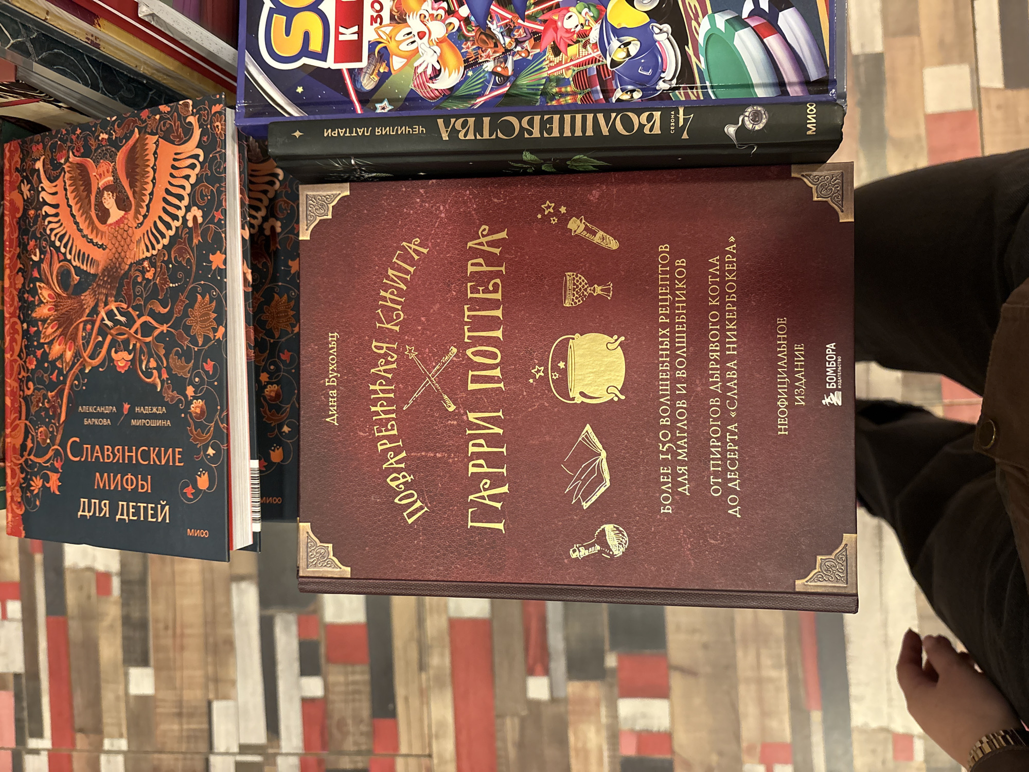 The image shows a stack of books. On the bottom is a cookbook titled "Harry Potter Cookbook" in Russian. Above it are books with titles "4 Wizardry Season" and "Slavic Myths for Children", also in Russian. A person's hand is visible in the lower right corner. The books are arranged on a surface with a colorful striped pattern. The city of Saint Petersburg, Russia, is not directly evident in the image, but the books being in Russian suggests it could be located in Russia or another Russian-speaking region.