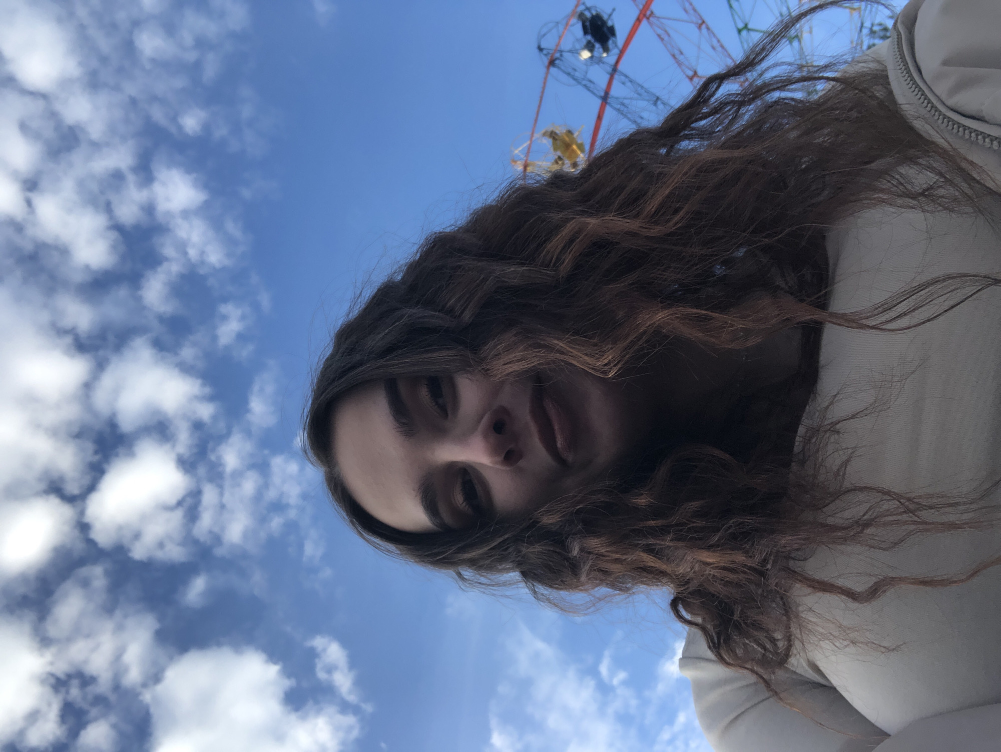 A low-angle photograph captures a young adult female, visible from the chest up, positioned against a clear blue sky with scattered white cumulus clouds. She possesses long, dark, wavy hair and looks directly at the camera with a slight smirk. She is wearing a light-colored, long-sleeved top or jacket, with a visible zipper on the lower left side of the frame. In the upper right background, a portion of a red-framed Ferris wheel is visible, featuring distinct yellow and dark-colored gondolas. This outdoor scene is situated within an amusement park or fairground in the city of Chaikovsky, Russia.